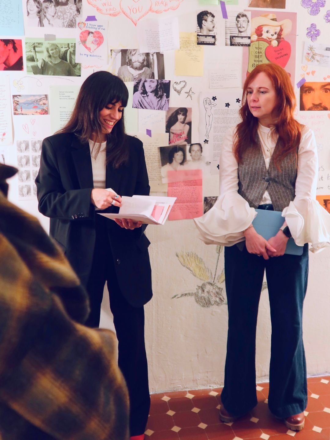 Momento di confronto e regia editoriale durante una mostra d'arte. Due professioniste analizzano i materiali cartacei e la documentazione visiva installata a parete.