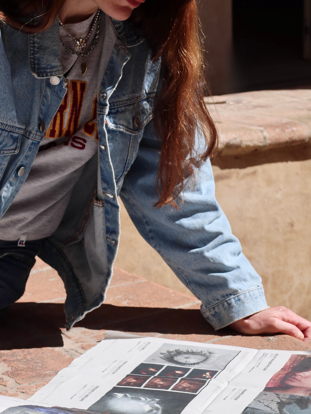 Una persona con giacca di jeans e una maglietta leggendo un giornale con immagini di scatti fotografici, seduta su una superficie di pietra.