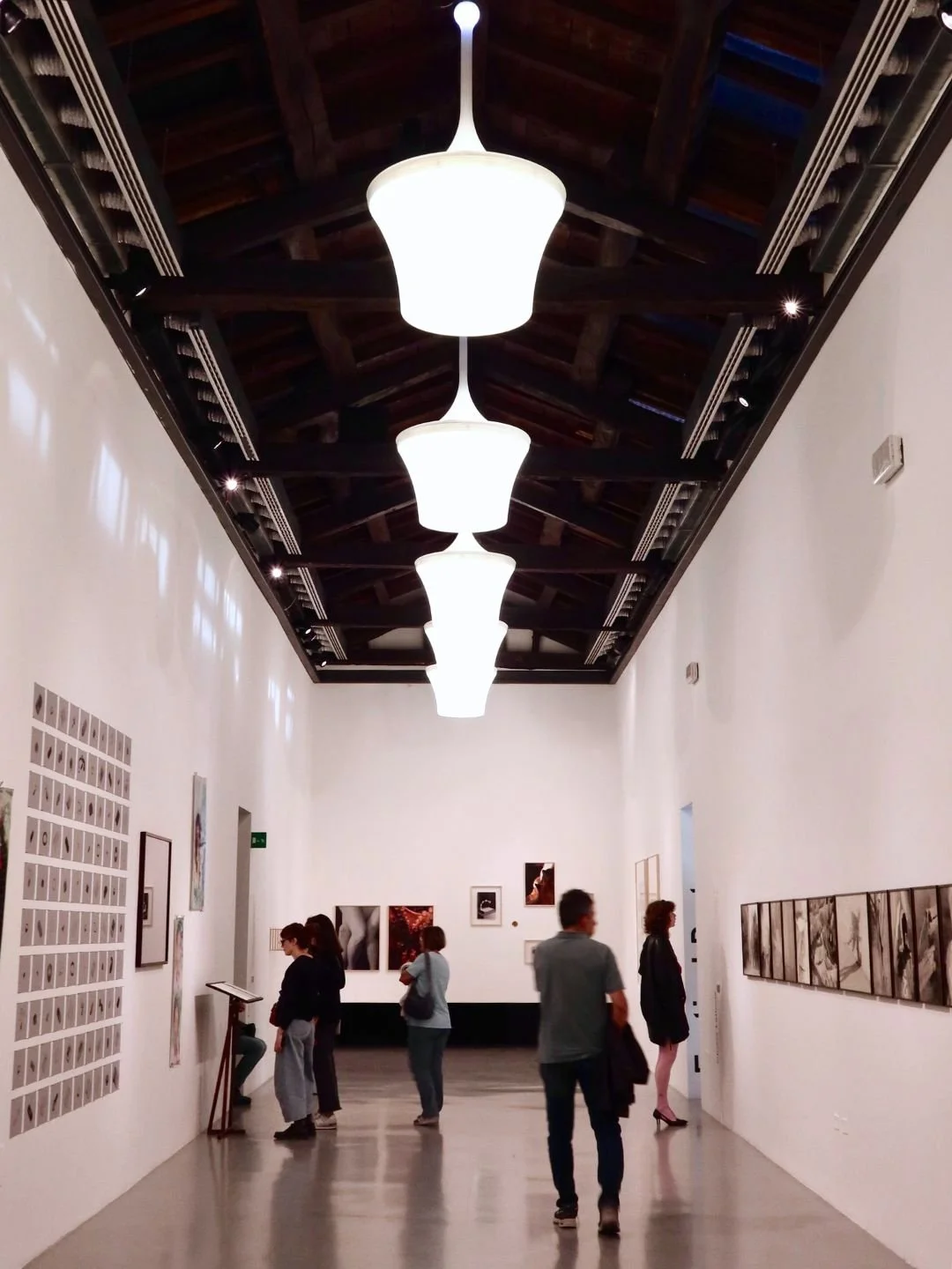 Sala di un museo con pareti bianche, diverse opere d'arte e persone che osservano le esposizioni, illuminata da grandi lampade a soffitto