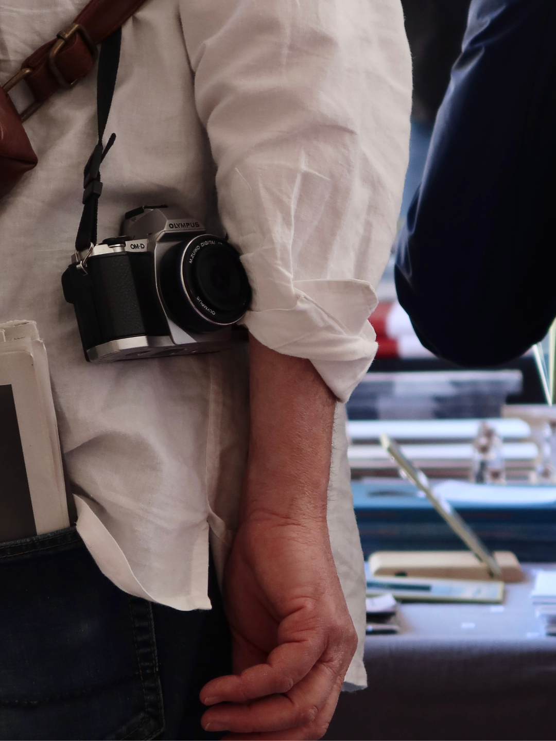 Persona con camicia bianca e macchina fotografica appesa al collo, con mano penzolante accanto a una libreria o espositore