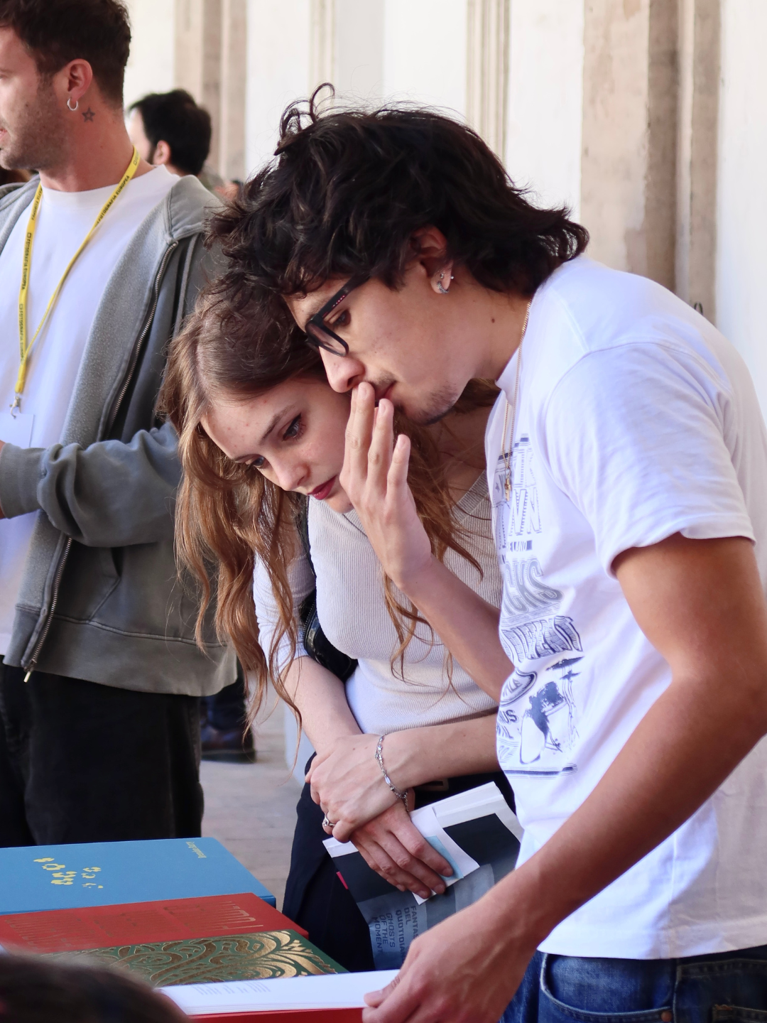 Due giovani persone stanno curiosando tra i libri su un tavolo durante un evento o una mostra, mentre alle loro spalle ci sono altre persone.