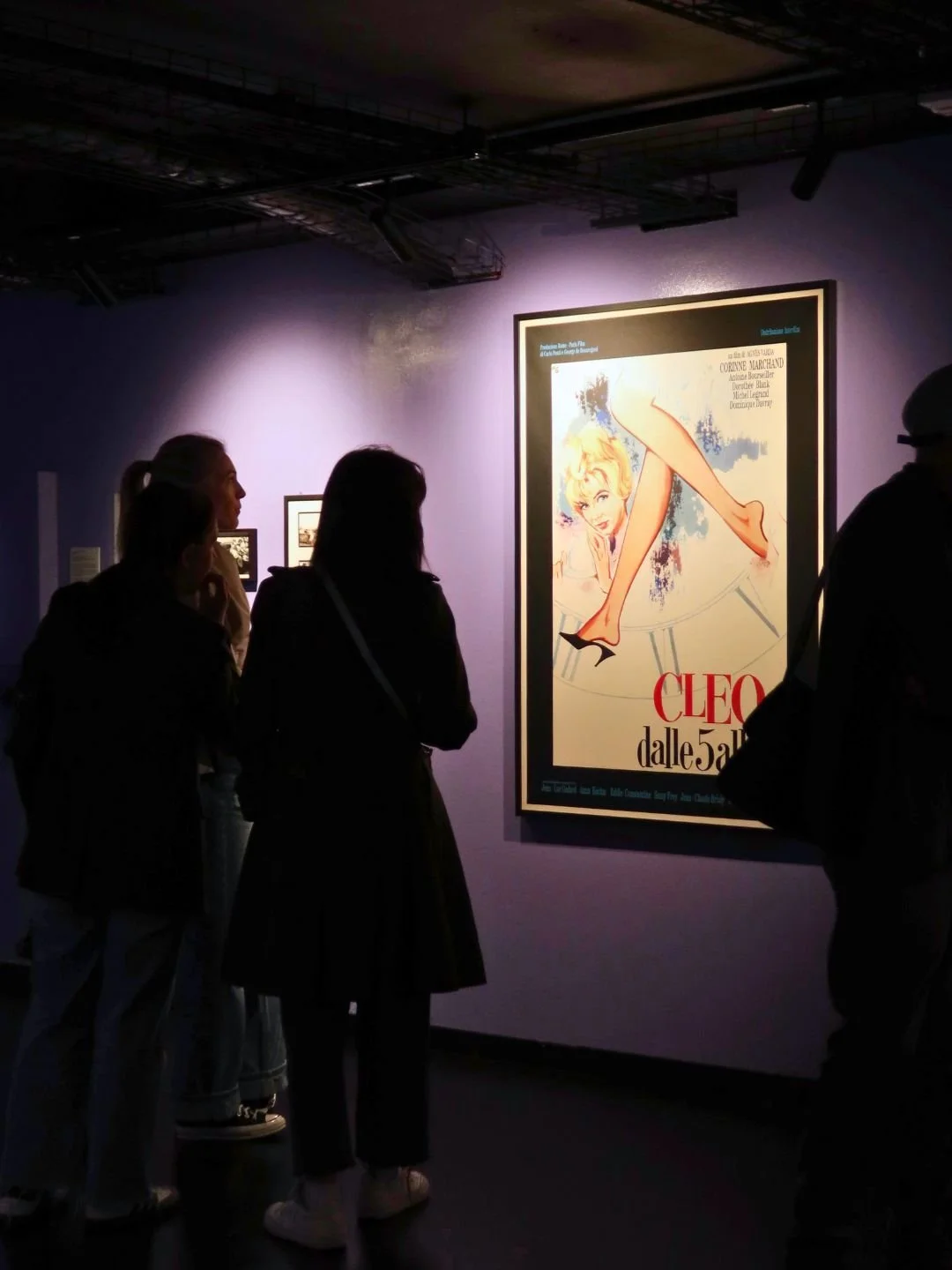 Gente che guarda un poster di un film, uno stile vintage, con un personaggio femminile decorato con un paio di gambe lunghe e scarpe eleganti, in un ambiente di museo o galleria.