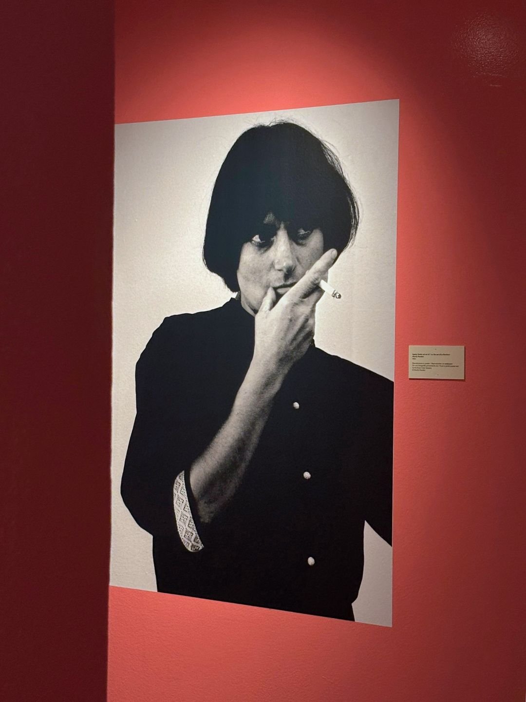 Fotografia in bianco e nero di una donna con capelli corti, vestita di nero, che fuma una sigaretta. La donna ha un'epoca degli anni '60 o '70 e si trova in un'esposizione artistica su uno sfondo bianco e rosa.