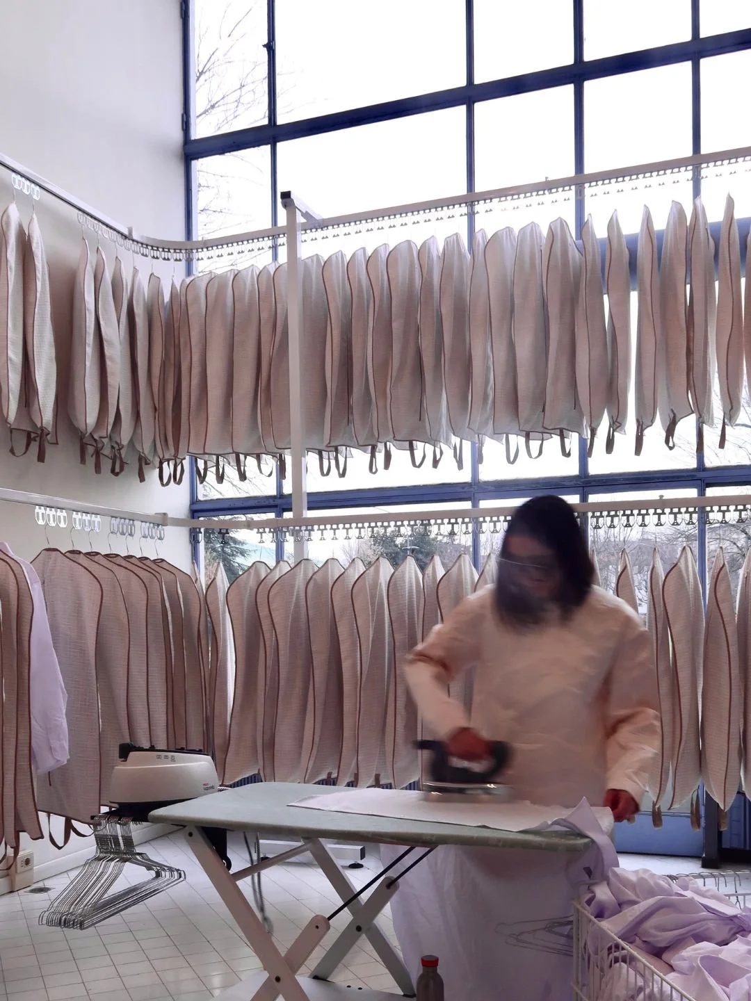 Lavoratrice che stira tessuti in un laboratorio con tende di tessuto appese e grandi finestre.