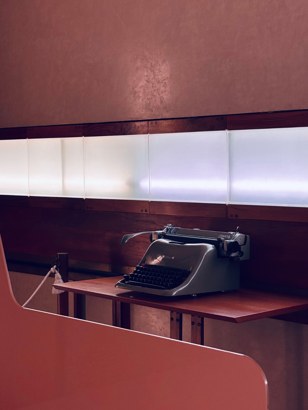 Una macchina da scrivere vintage di colore grigio posizionata su un tavolo di legno, con una parete di pannelli luminosi e una parete di legno sullo sfondo.