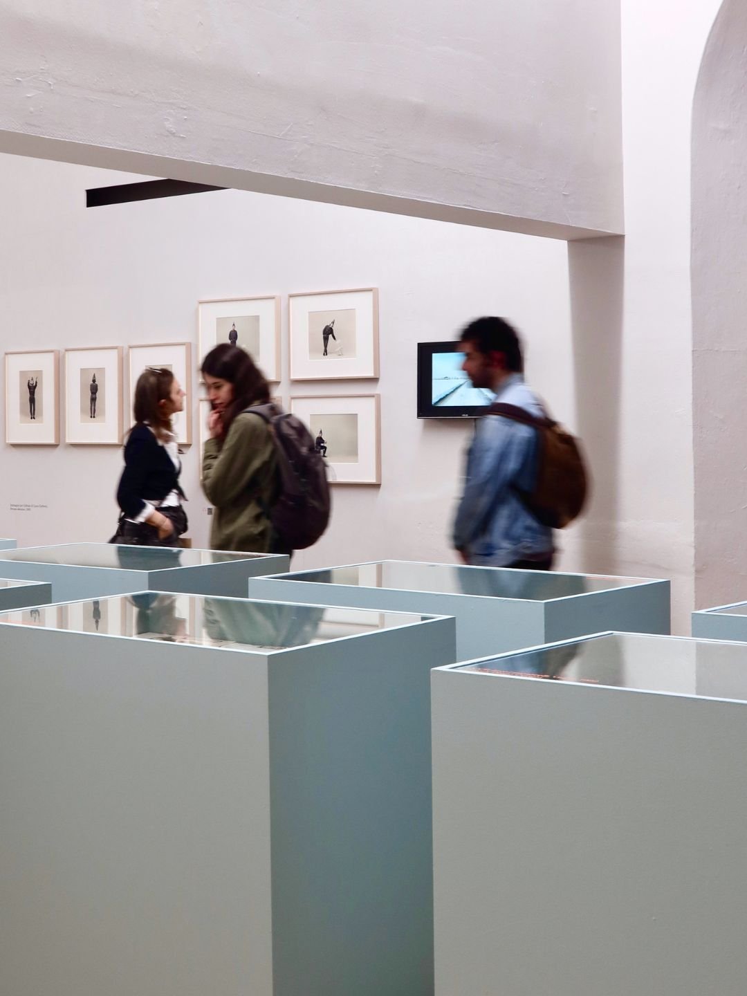 Visitatori in un museo d'arte contemporanea, con fotografie di figure umane appese alle pareti e esposizioni di vetro in primo piano.