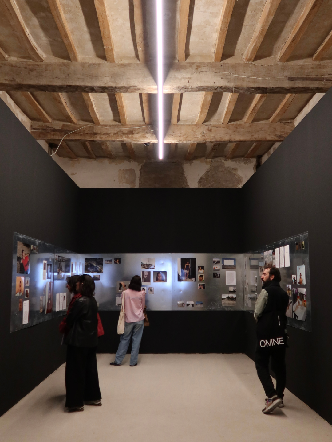Visitatori in una galleria d'arte con pareti nere e postazioni di foto e testo, soffitto in legno con travi esposte e illuminazione moderna.