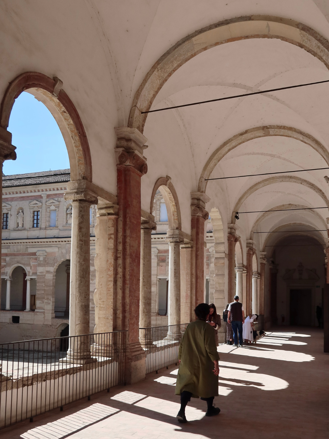 Portico storico con archi e colonne in pietra, persone che camminano sotto le arcate, facciata di un edificio storico visibile all'esterno, luce solare che crea ombre sul pavimento.
