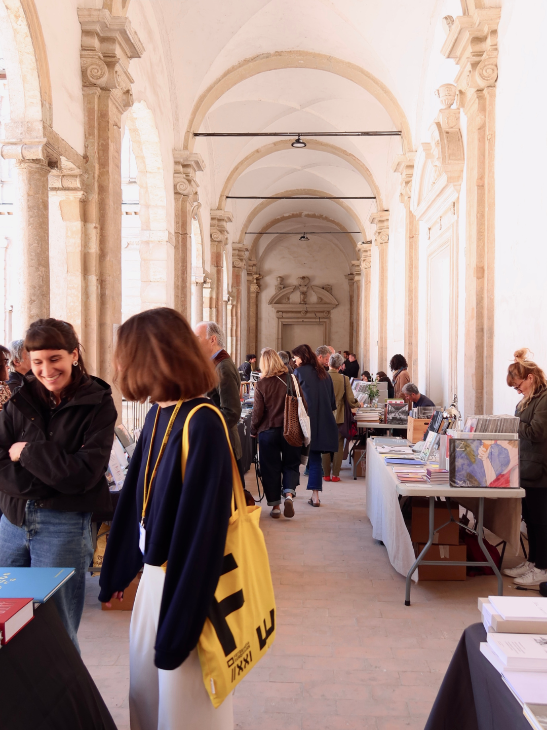 Folla di persone che visitano una fiera del libro in un edificio storico con archi e colonne in pietra.