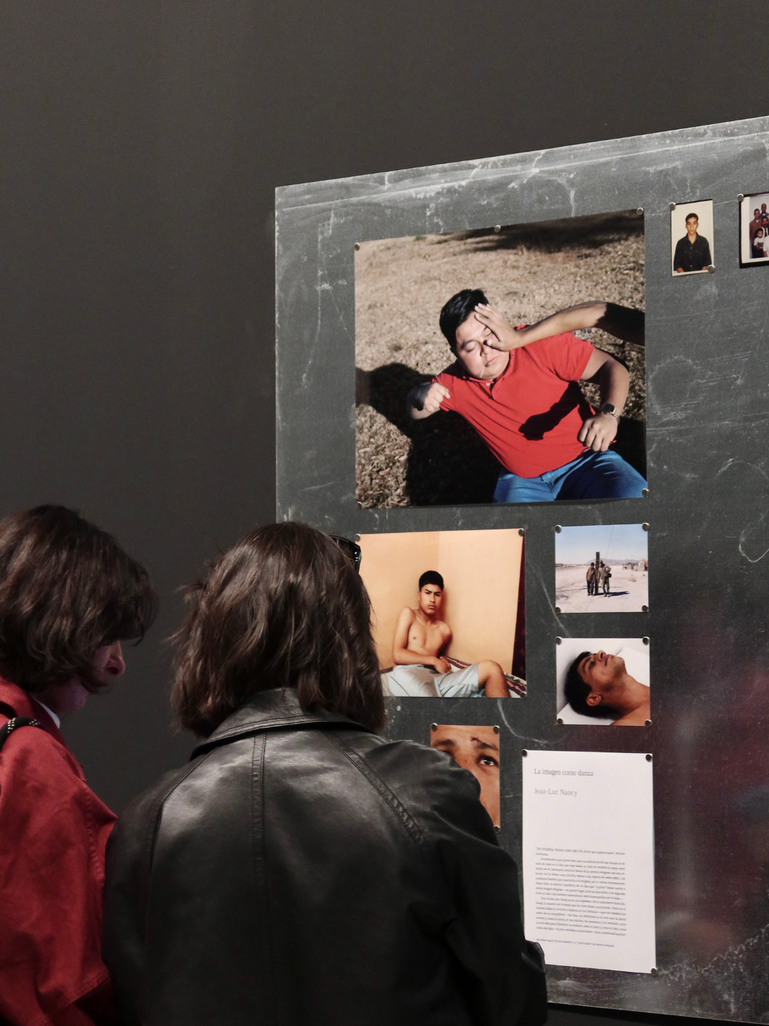 Due persone osservano una parete con fotografie: una di un uomo con gli occhi chiusi, una di un ragazzo senza maglietta seduto, e altre immagini di uomini in ambienti naturali.