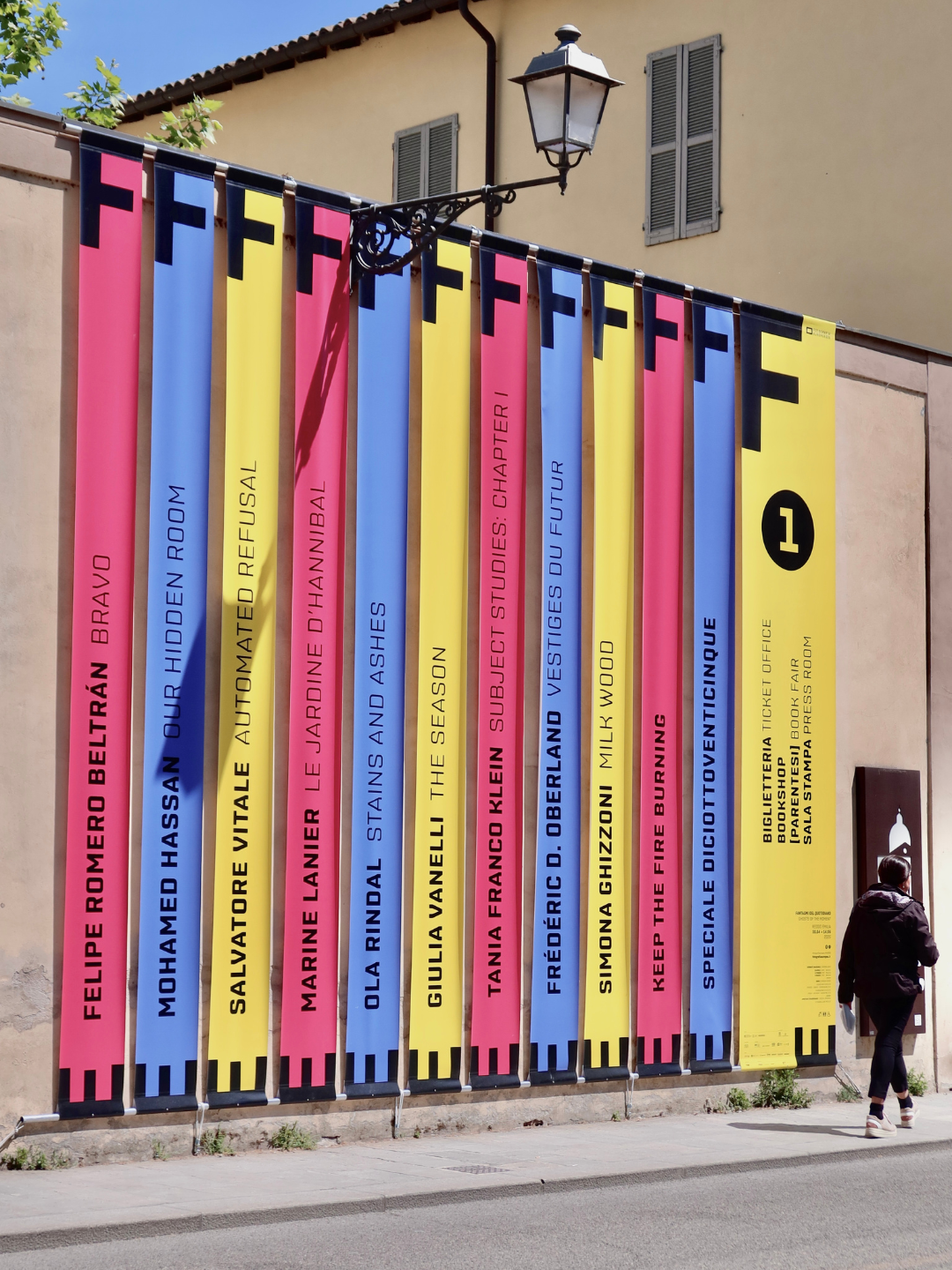 Bandiere colorate con titoli di libri e autori in italiano e inglese, appese a una corda lungo una strada davanti a un edificio.