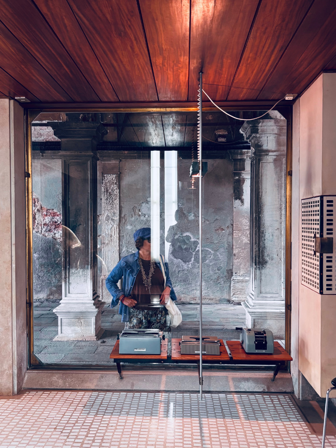 Donna con cappello blu, occhiali e una borsa, che osserva vari dispositivi tecnologici esposti dietro una vetrina in un ambiente storico con colonne e pareti vecchie.
