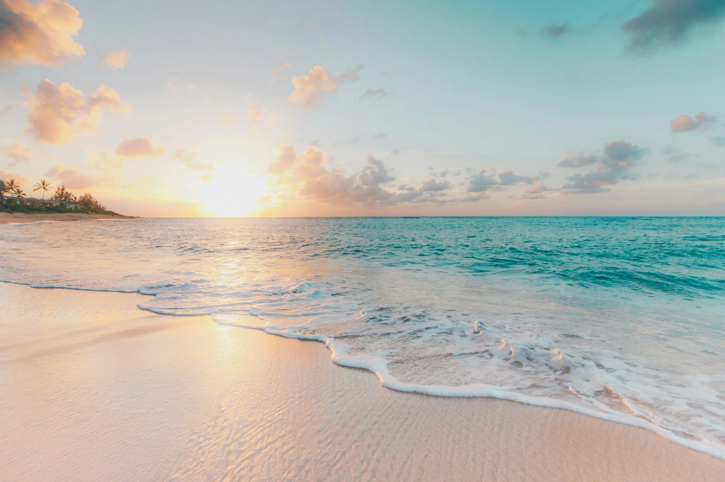 A serene beach scene during sunset with gentle waves washing onto the sandy shore, palm trees on the left, and a sky filled with soft clouds illuminated by the setting sun.