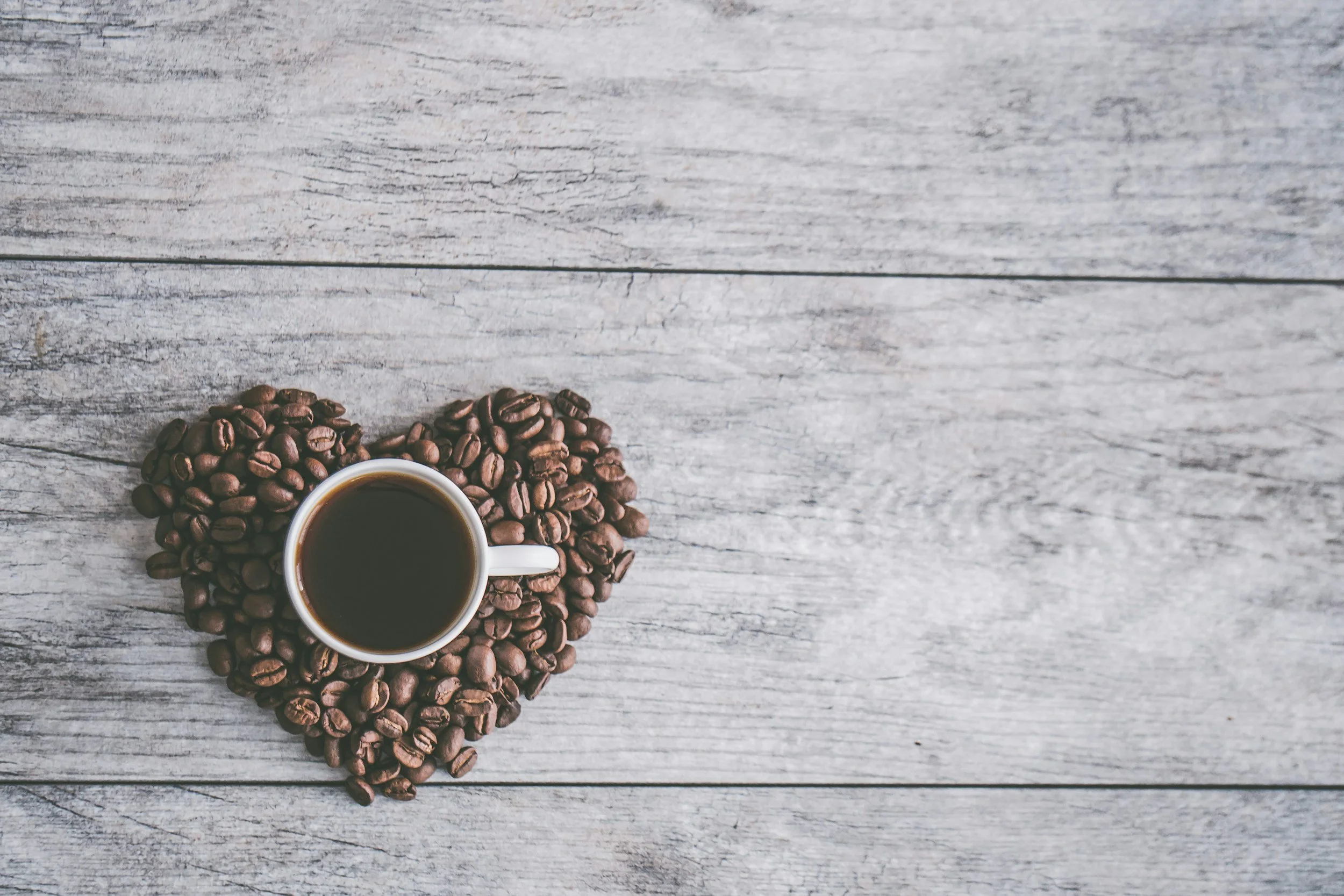 A white coffee cup filled with black coffee placed on a heart-shaped arrangement of coffee beans on a wooden surface.
