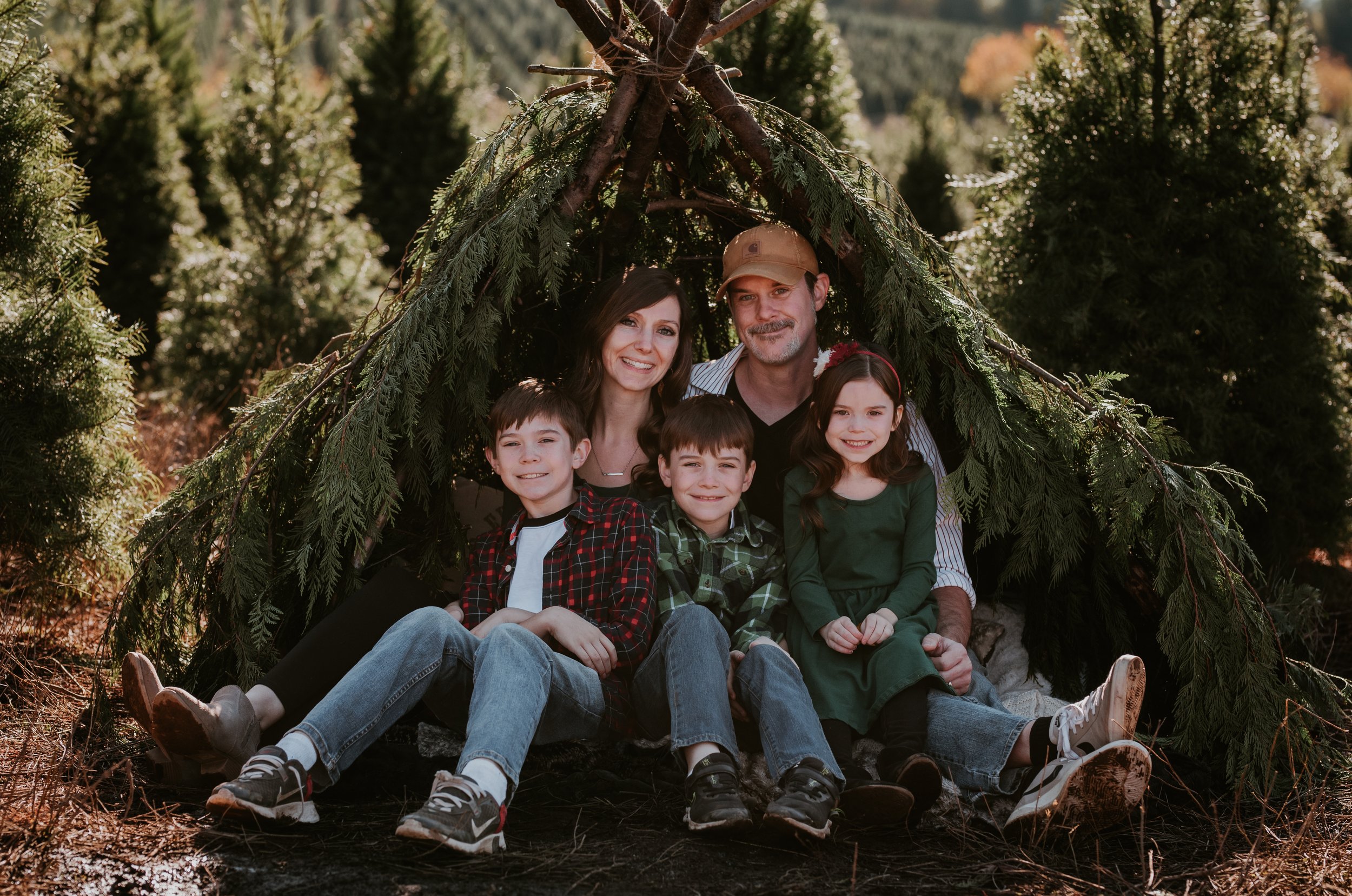 A family of five sitting under a makeshift shelter made of tree branches and greenery outdoors.