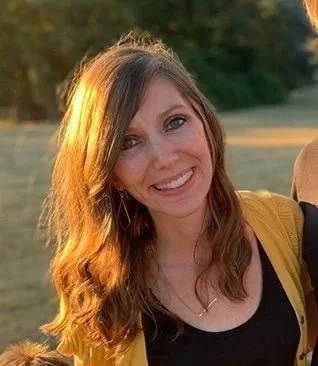 A woman with long, wavy brown hair smiling outdoors during sunset, wearing a black top and a yellow jacket, with a grassy field and trees in the background.