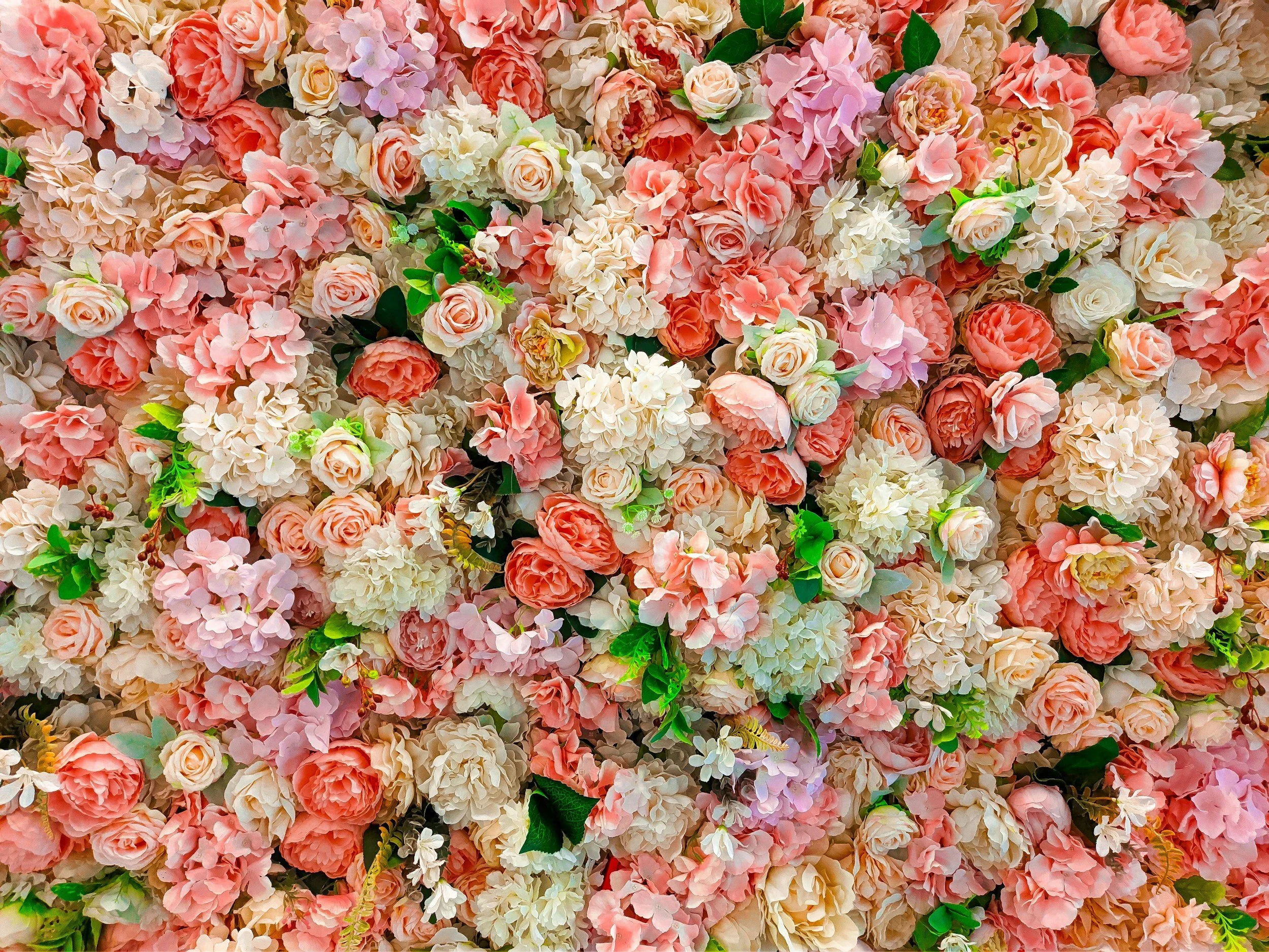 A dense arrangement of pink, white, and peach roses, hydrangeas, and other flowers with green leaves, creating a floral backdrop.