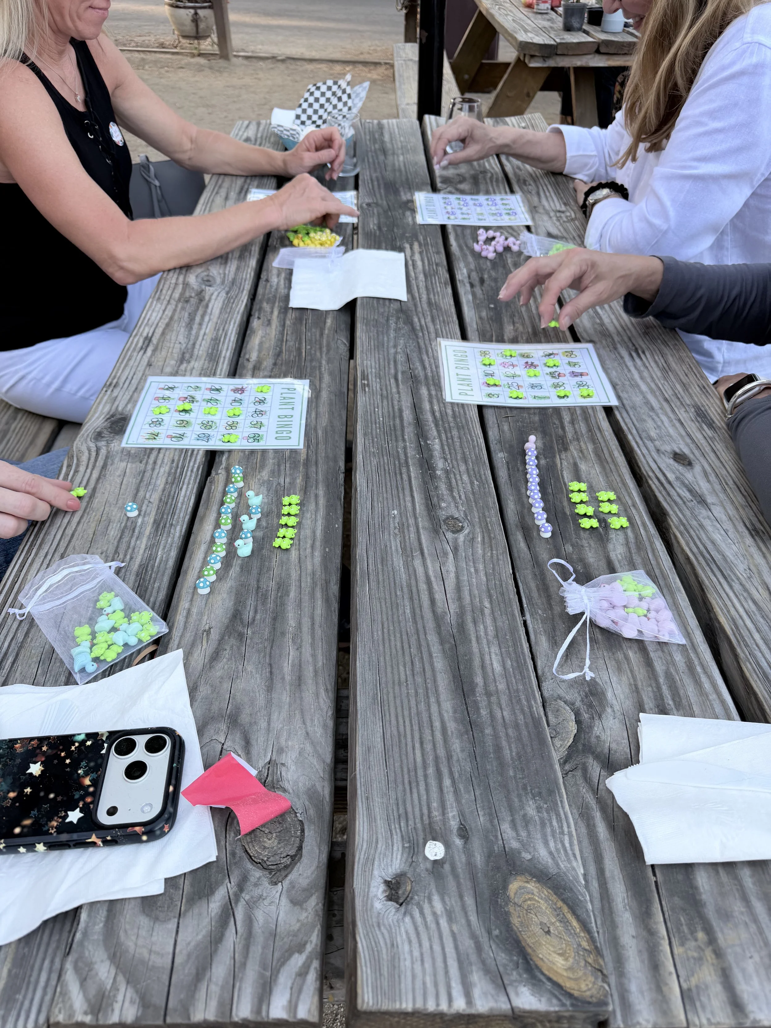 People playing a bingo game at an outdoor wooden table, with bingo cards, small colorful game pieces, and some personal belongings on the table.