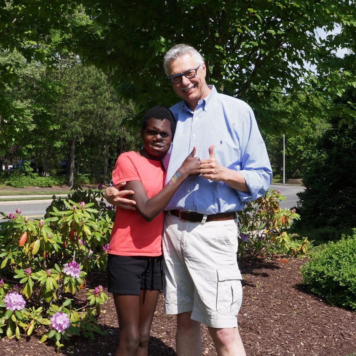 A young girl and an older man standing outdoors in front of green trees and bushes, embracing and smiling at the camera.