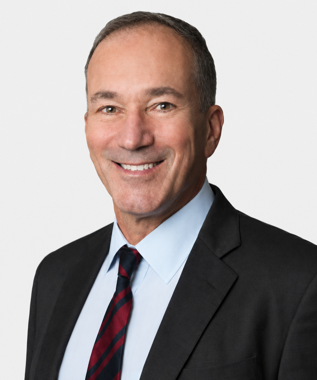 A professionally dressed man with short brown hair smiling at the camera. He is wearing a black suit jacket, light blue shirt, and a red and navy striped tie against a white background.