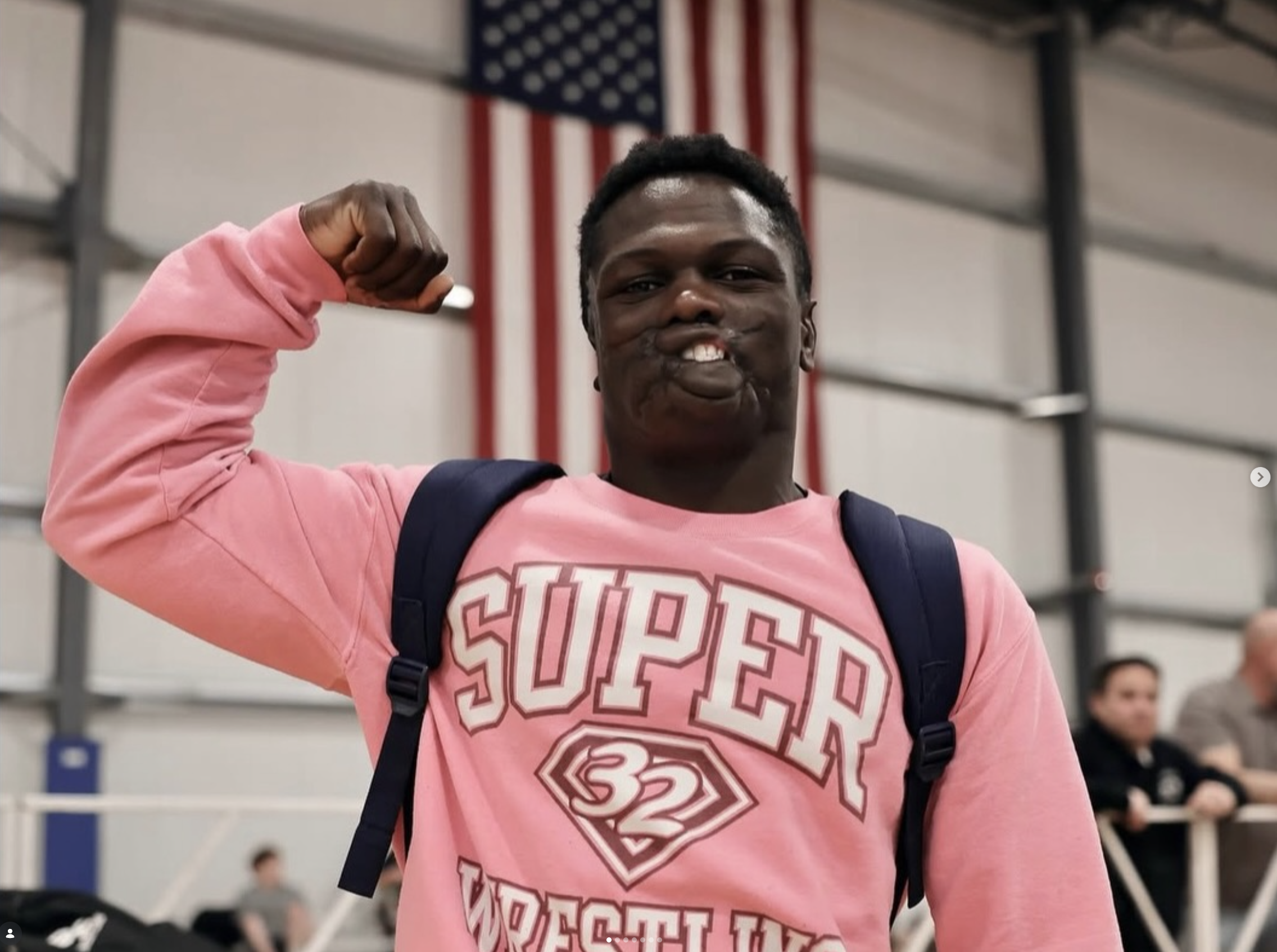 Dunia Sibomana-Rodreiguez is flexing his arm in a gym with an American flag in the background, wearing a pink hoodie and carrying a backpack.