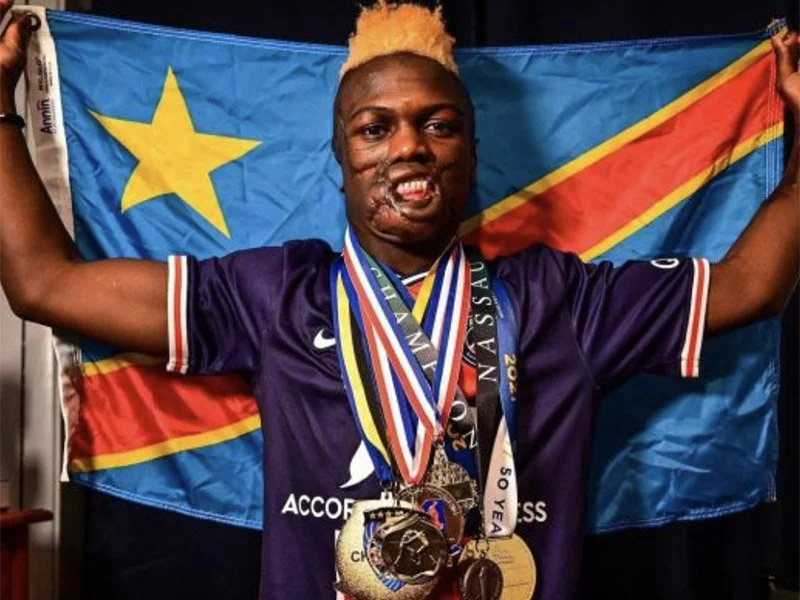 Person celebrating with medals and a flag of the Democratic Republic of the Congo, wearing a purple sports jersey, smiling with arms raised.