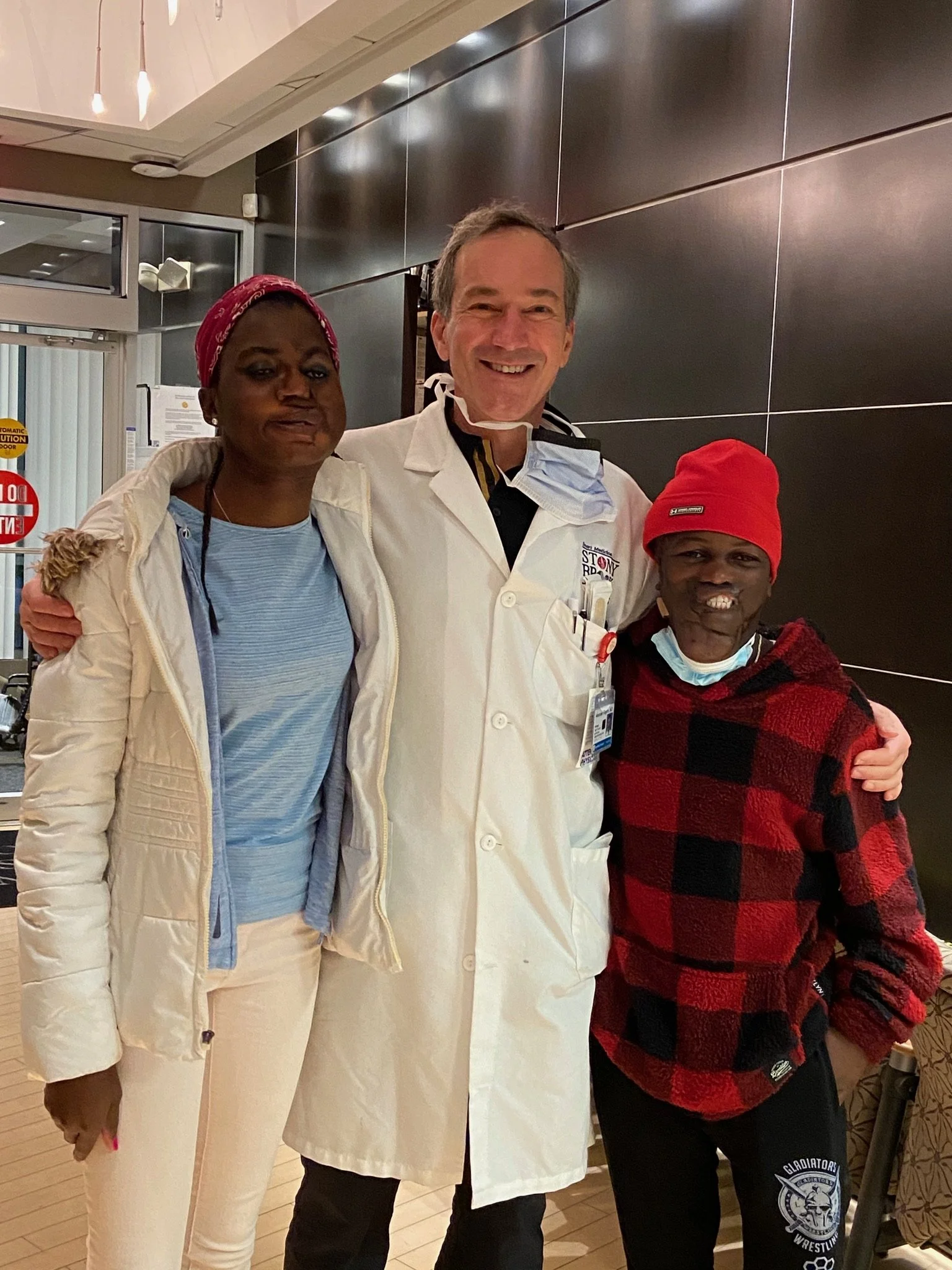 Three people standing together indoors; a woman on the left, a man in the middle wearing a white coat, and a person on the right in a red hat and red and black checkered jacket.