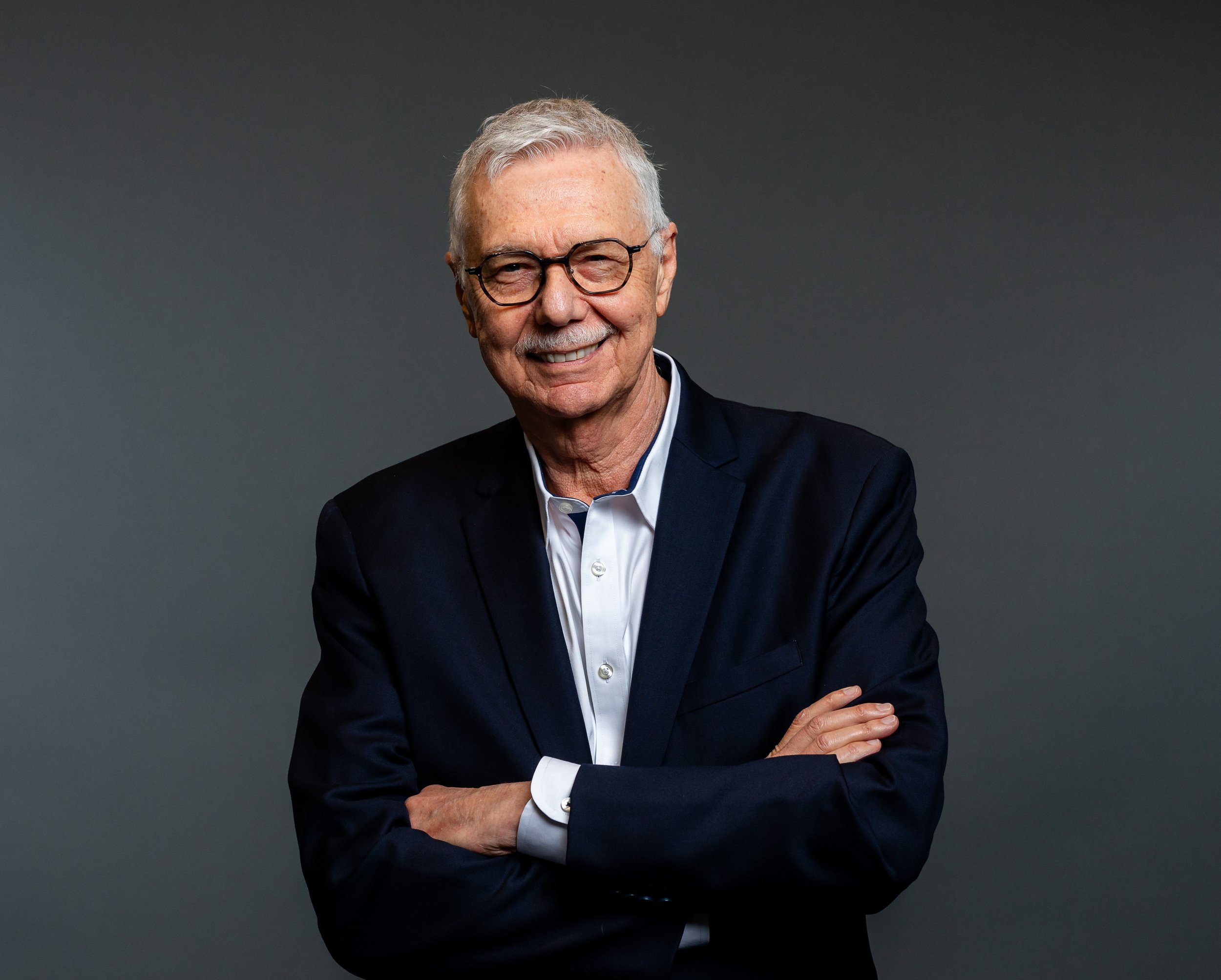 Dr. Leon Klempner, who has grey hair and glasses, is dressed in a dark suit and white shirt, and is smiling and standing with his arms crossed against a dark gray background.