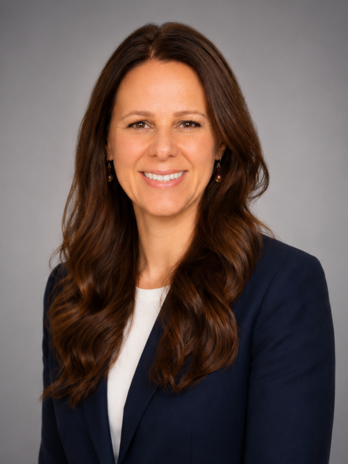 A woman with long brown wavy hair, smiling, wearing a navy blazer and white top, against a gray background.