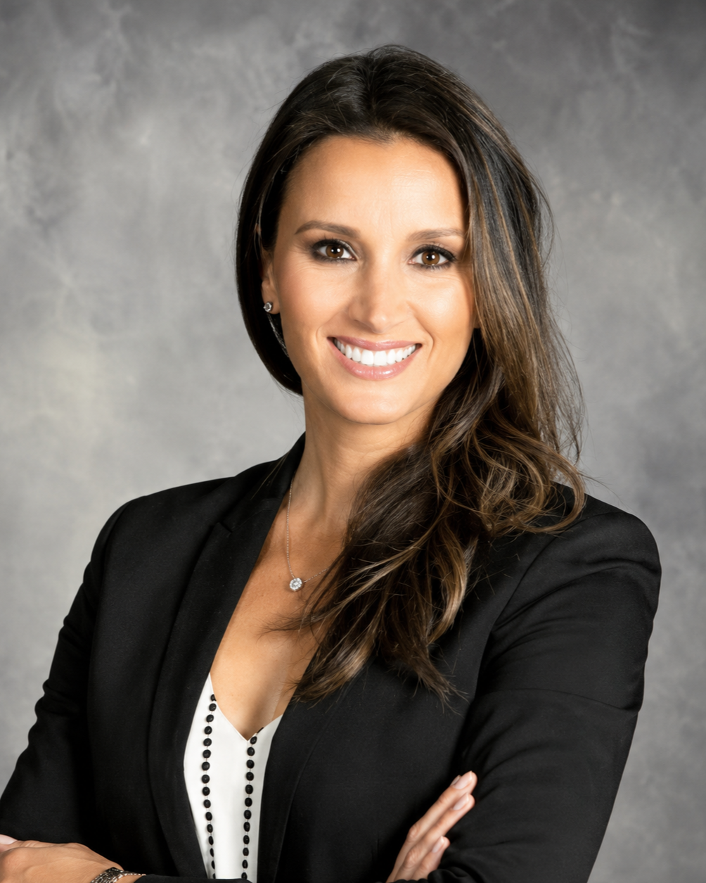 Professional woman with long brown hair smiling, wearing a black blazer, white blouse, and silver jewelry, with arms crossed in front of a gray background.