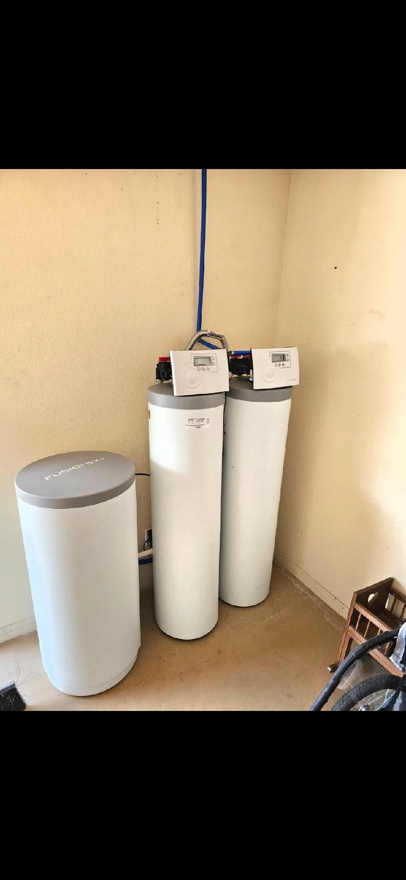 Water softener system with two large vertical cylinders and a control panel on top, situated in a corner of a room.