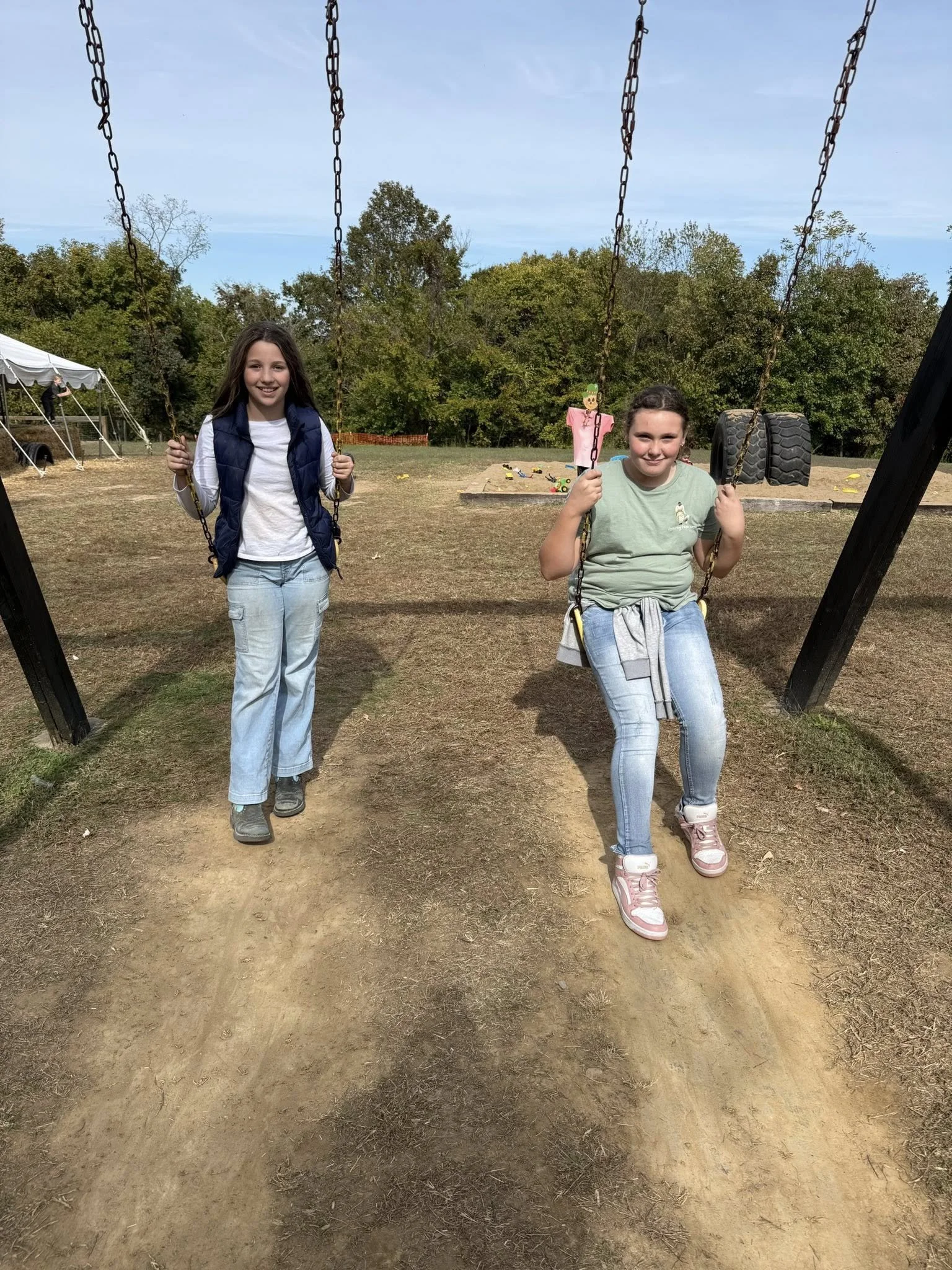 Two young girls at a park, one standing with swing chains, the other sitting on a swing, with trees, a tent, and large tires in the background on a partly cloudy day.