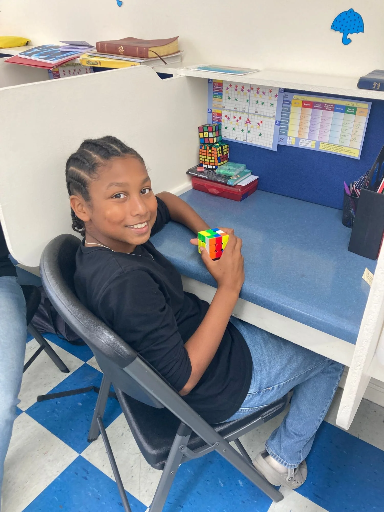 A young boy with braided hair sitting at a desk holding a colorful Rubik's Cube, smiling at the camera in a classroom or study area.