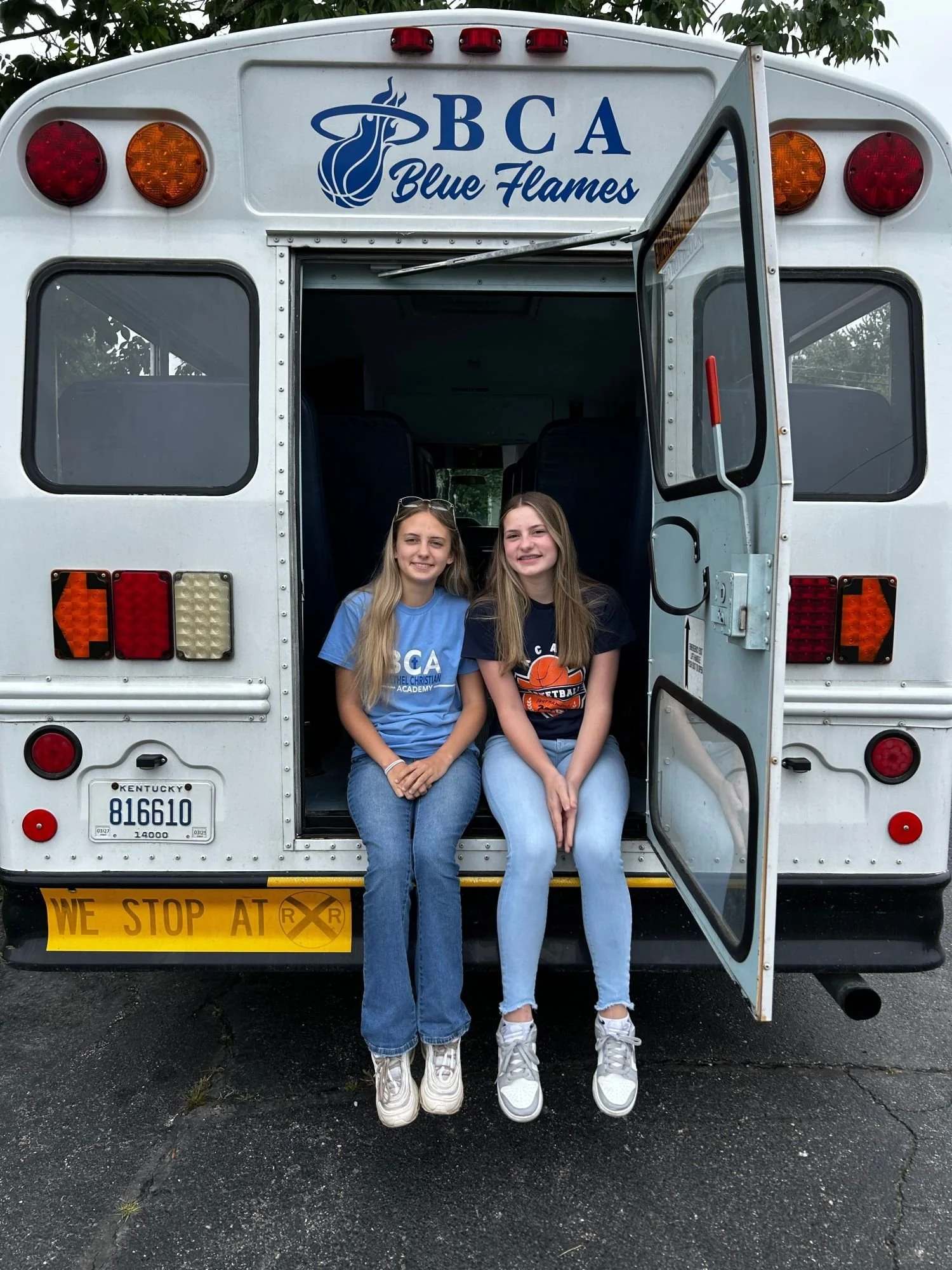 Two young girls are sitting at the back of a white school bus with the door open. The bus has the logo and text "BCA Blue Flames" on it. The girls are smiling and wearing T-shirts, one with a basketball logo. They are wearing sneakers and sitting on a black asphalt surface.