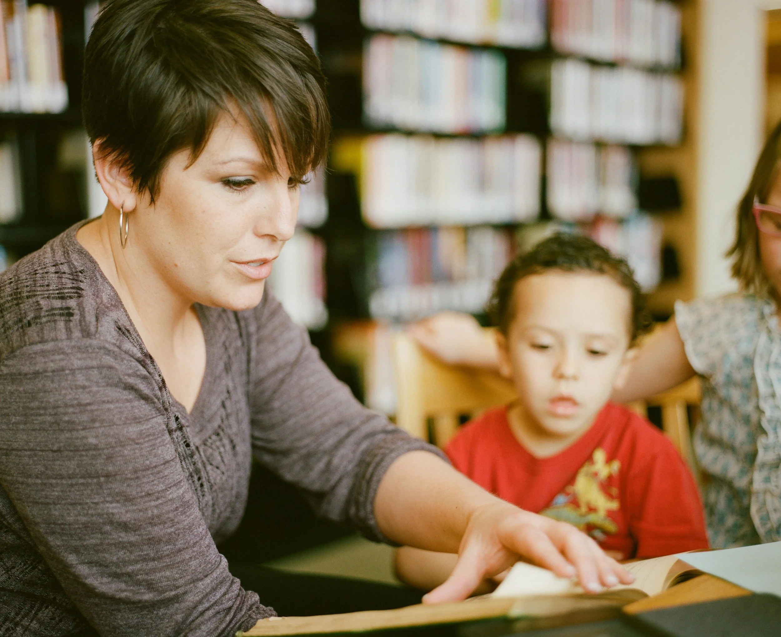 A woman with short dark hair wearing hoop earrings reads a book with a young boy in a red shirt at a table in a library.