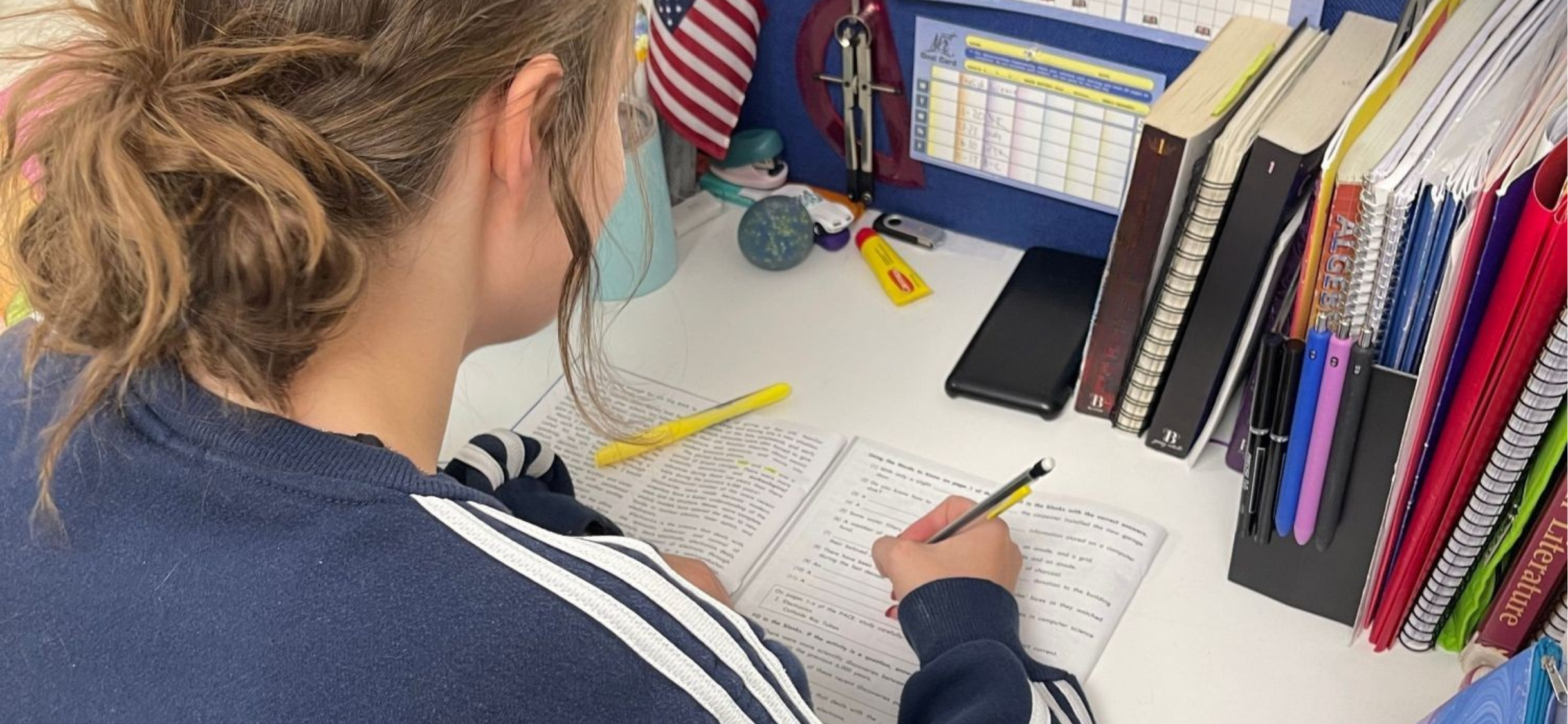 A student with curly hair, wearing a navy blue sweater with white stripes, sitting at a school desk, writing in a notebook with a yellow pen. The desk has various stationery and a large collection of books and binders on the right side.