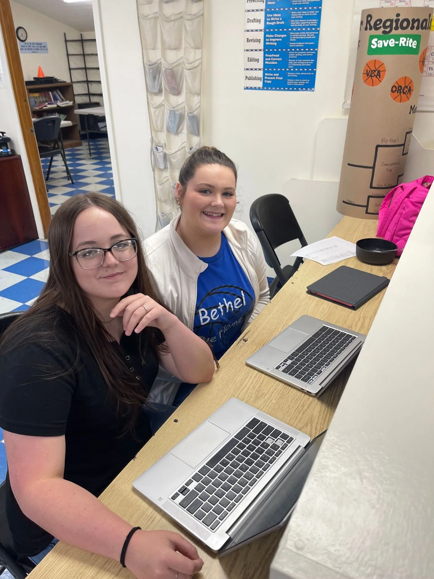 Two young women sitting at a wooden table in a classroom or library, smiling at the camera. One is wearing glasses and a black shirt, the other is wearing a white jacket and a blue shirt. On the table are two open laptops, papers, a black notebook, and a pink backpack. In the background, there is a white wall with a blue and white poster and a beige paper container with orange stickers. There is also a room with bookshelves, a table, and chairs visible through a doorway.