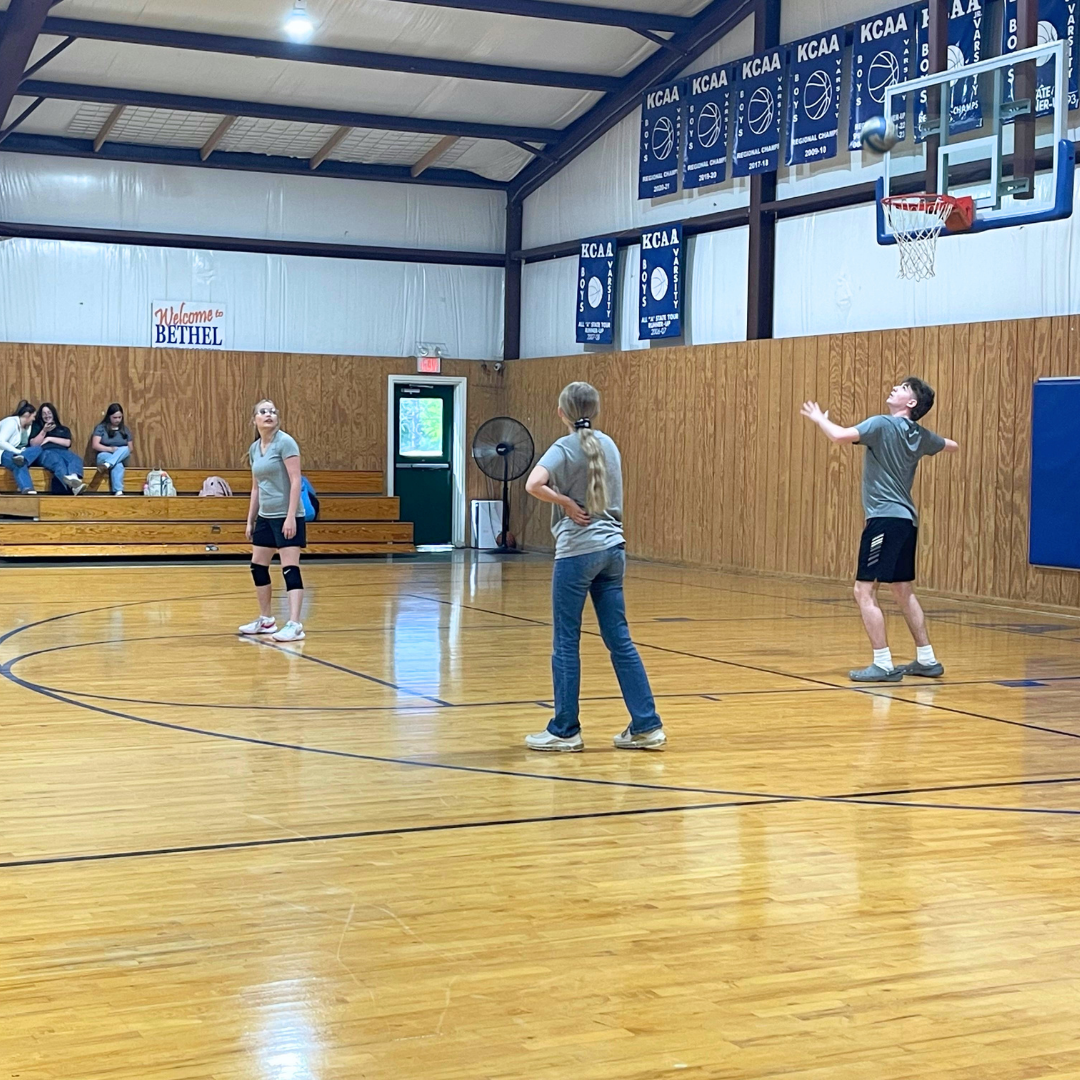 A girl in athletic attire and knee pads standing on a basketball court, watching a boy serve or shoot a basketball. A girl with long hair and a ponytail, wearing a gray T-shirt and jeans, stands with her hand on her hip. Several girls sit on the bleachers watching, with backpacks visible. The gym has wooden floors, banners hanging on the wall with sports achievement years, and a sign that says 'Welcome to Bethel'.