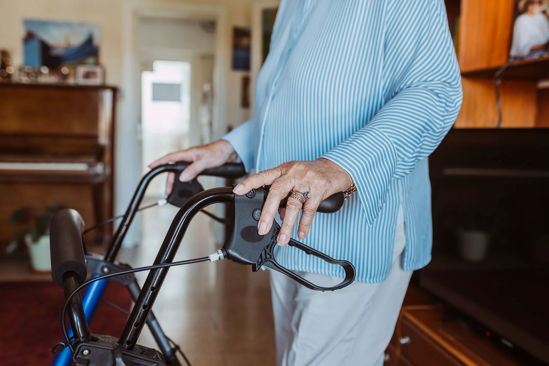 An elderly woman wearing a blue and white striped blouse using a walker with ergonomic handles inside a home with wooden furniture in the background.
