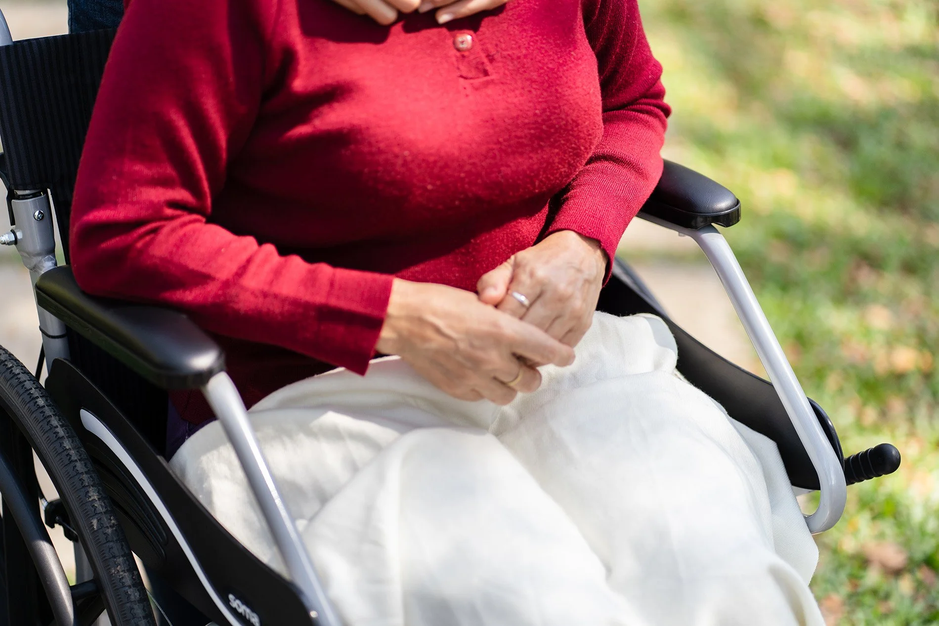 Close-up of a person sitting in a wheelchair outdoors, wearing a red sweater and beige pants, with hands resting on their lap, in a park or green area.