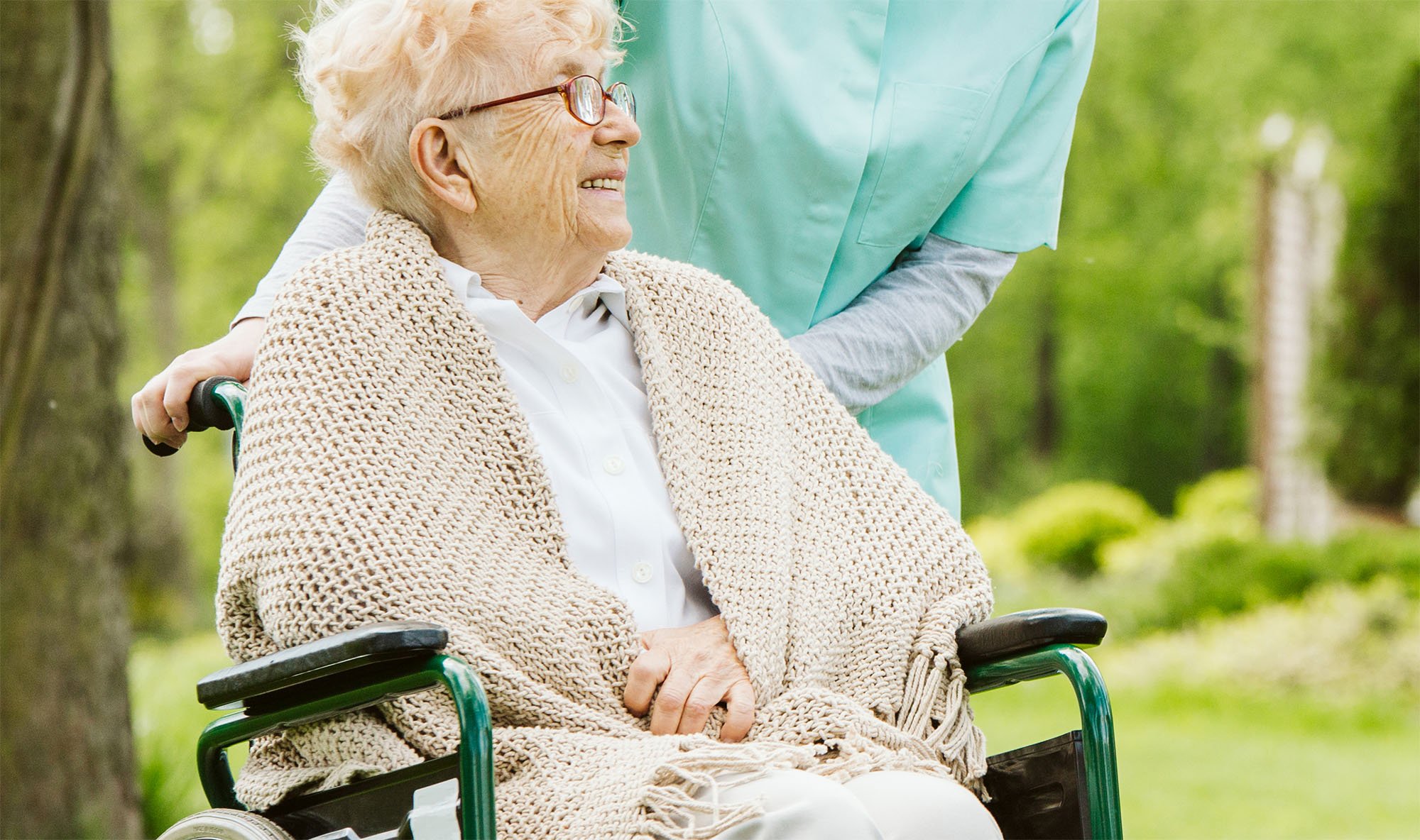 An elderly woman with glasses smiling while sitting in a wheelchair outdoors, with a caregiver standing behind her in a green park.