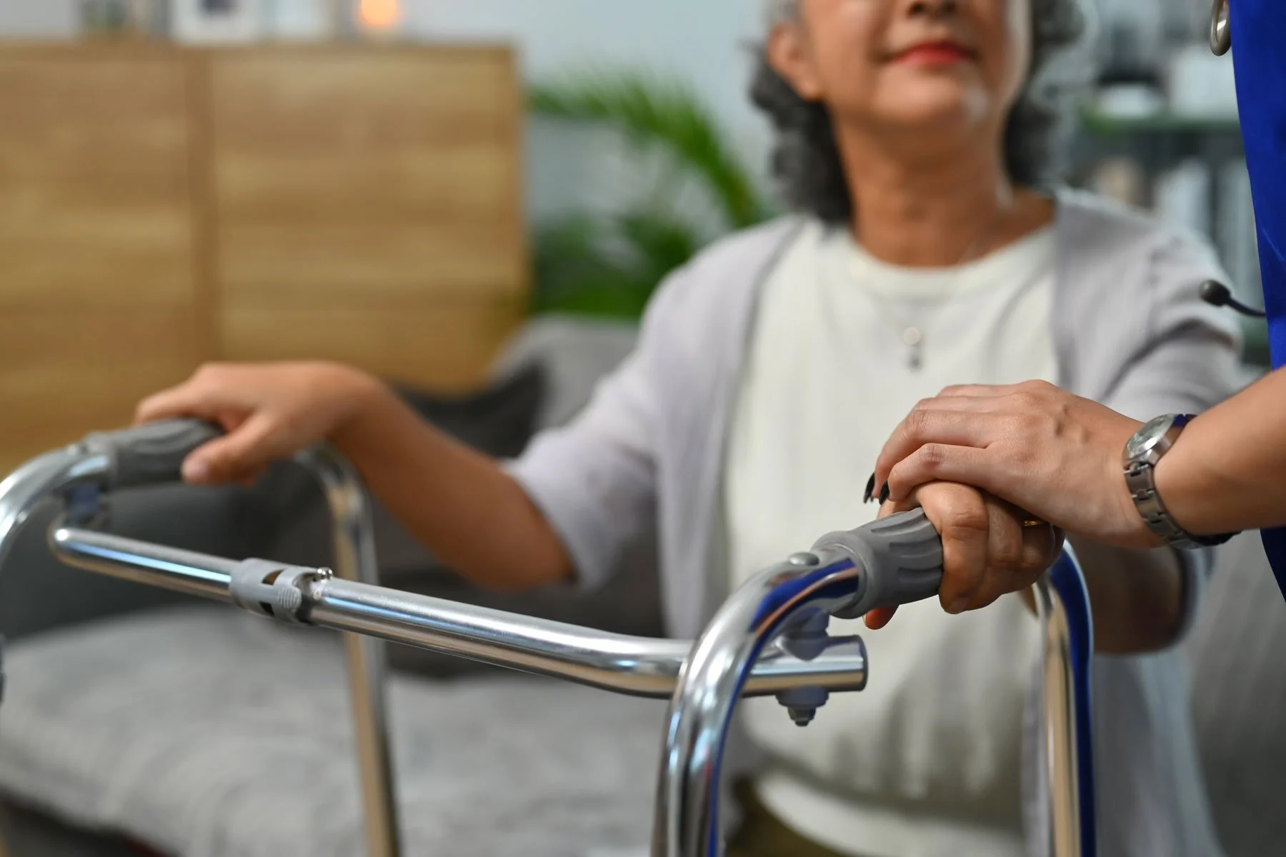 A woman in white clothing with a necklace in a wheelchair holding hands with a healthcare worker wearing a blue uniform and a watch, in a healthcare setting.