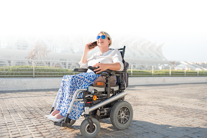 A smiling woman sitting in a motorized wheelchair outdoors in front of a modern architectural building, wearing sunglasses, a white top, and blue patterned pants.
