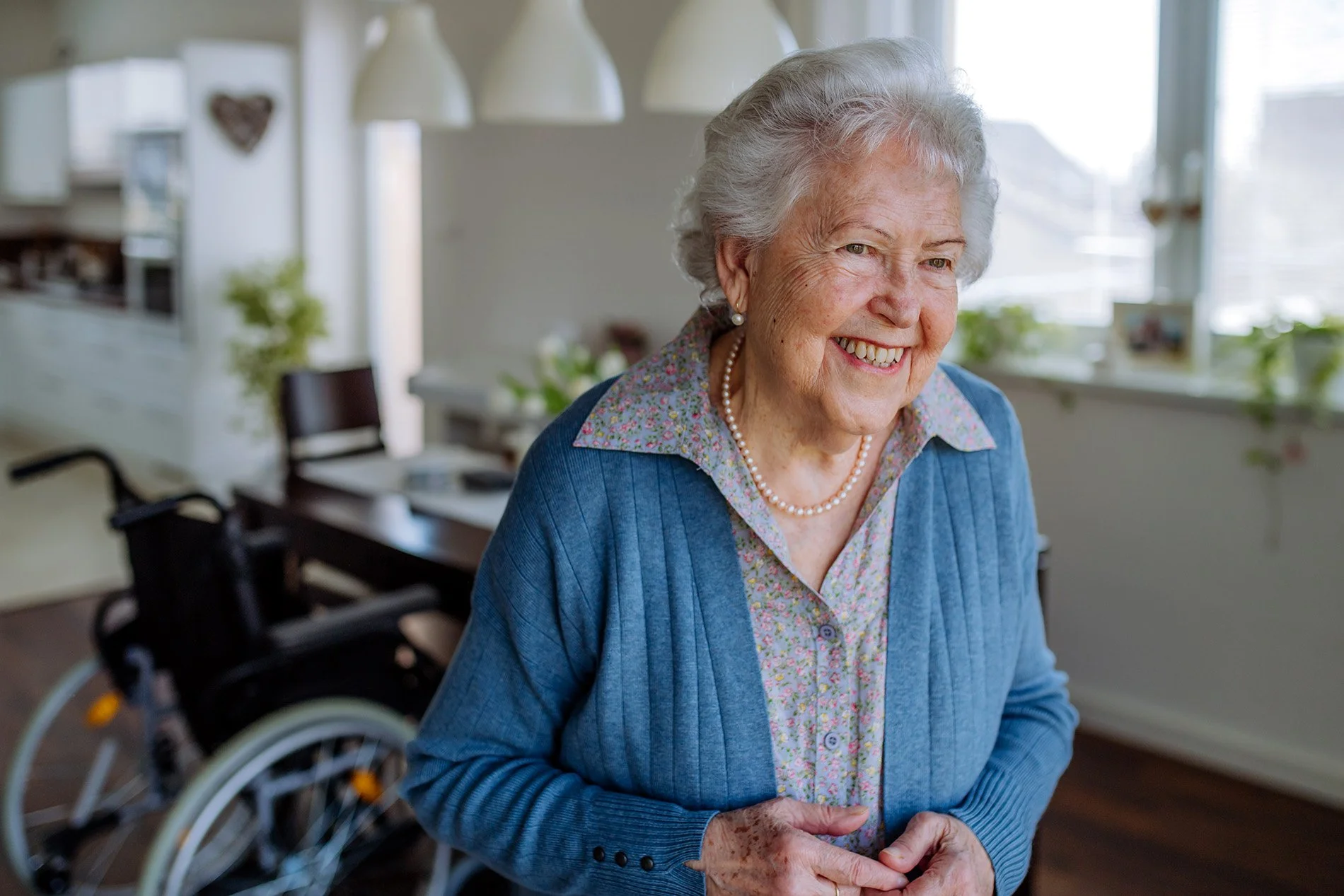 An elderly woman with gray hair smiling, wearing a floral blouse, pearl necklace, and a blue cardigan, in a bright room with windows and houseplants, with a wheelchair in the background.
