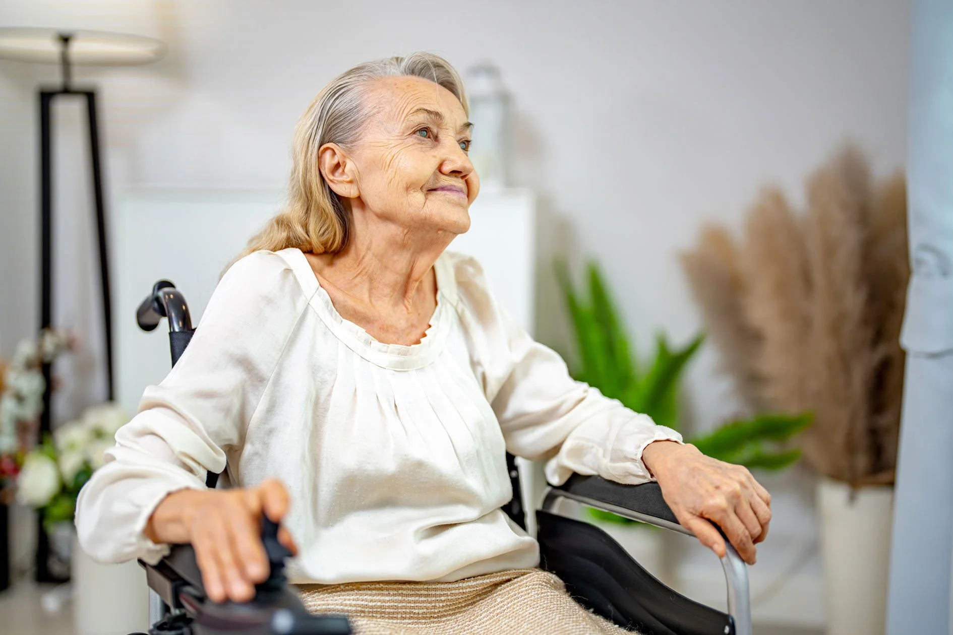 An elderly woman with gray hair and a warm expression sitting in a wheelchair in a bright room, surrounded by plants and soft decor.