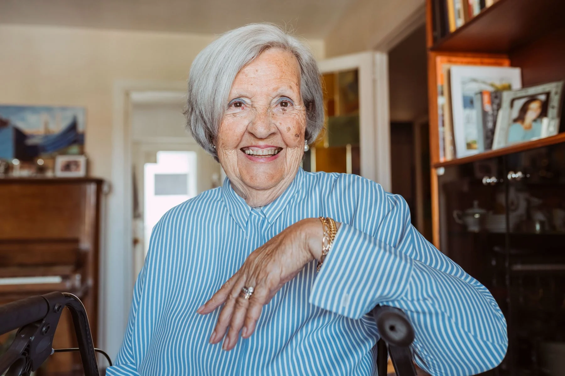 An elderly woman with gray hair smiling and wearing a blue and white striped shirt, sitting in a room with bookshelves and a piano, with her hand resting on a walker.