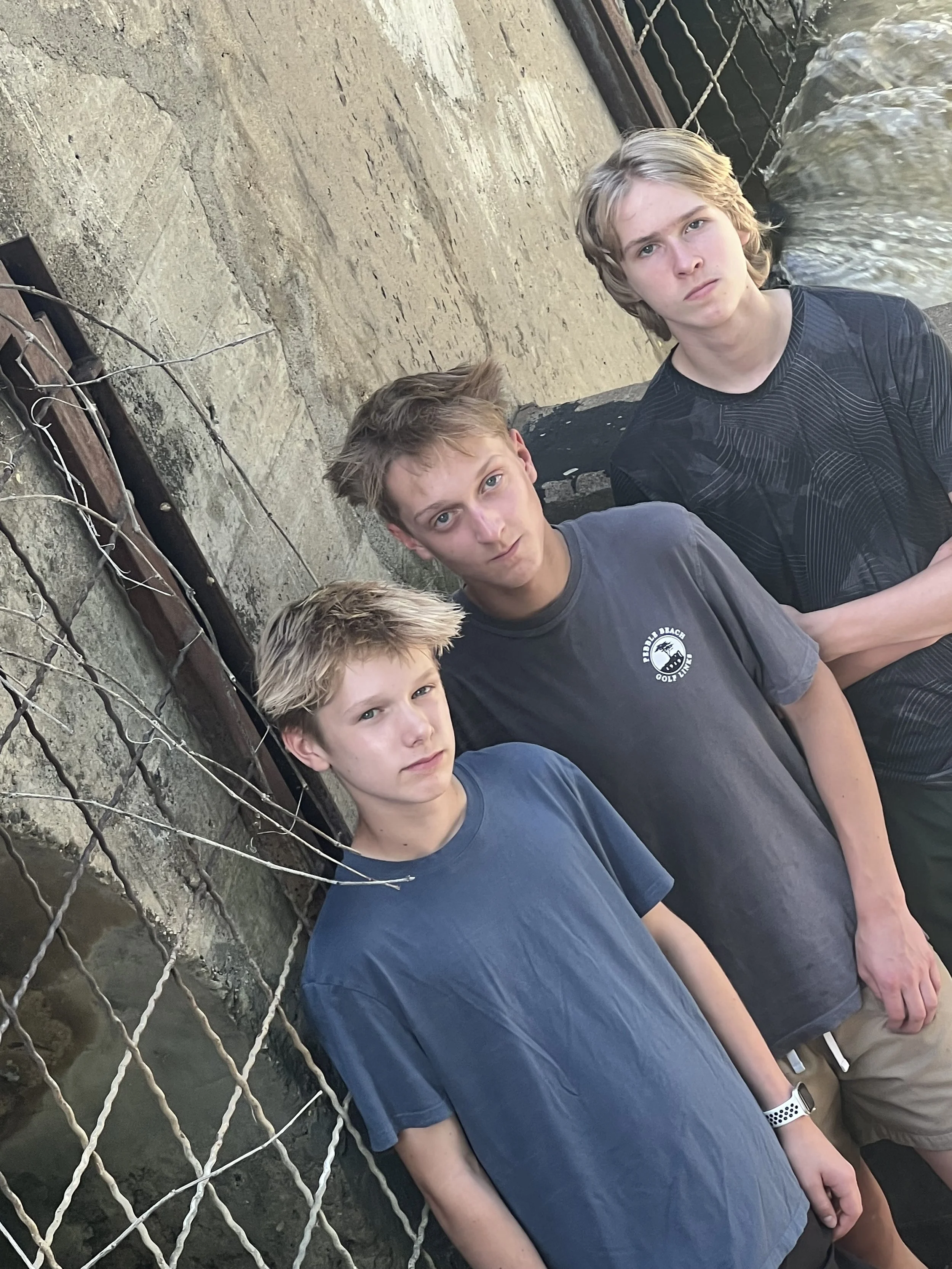 Four boys stand against a stone wall with a chain-link fence in front of them. One boy wears a blue T-shirt, another a gray T-shirt, and the others are in dark tops. They are outdoors near water, possibly at a waterfront or dock.