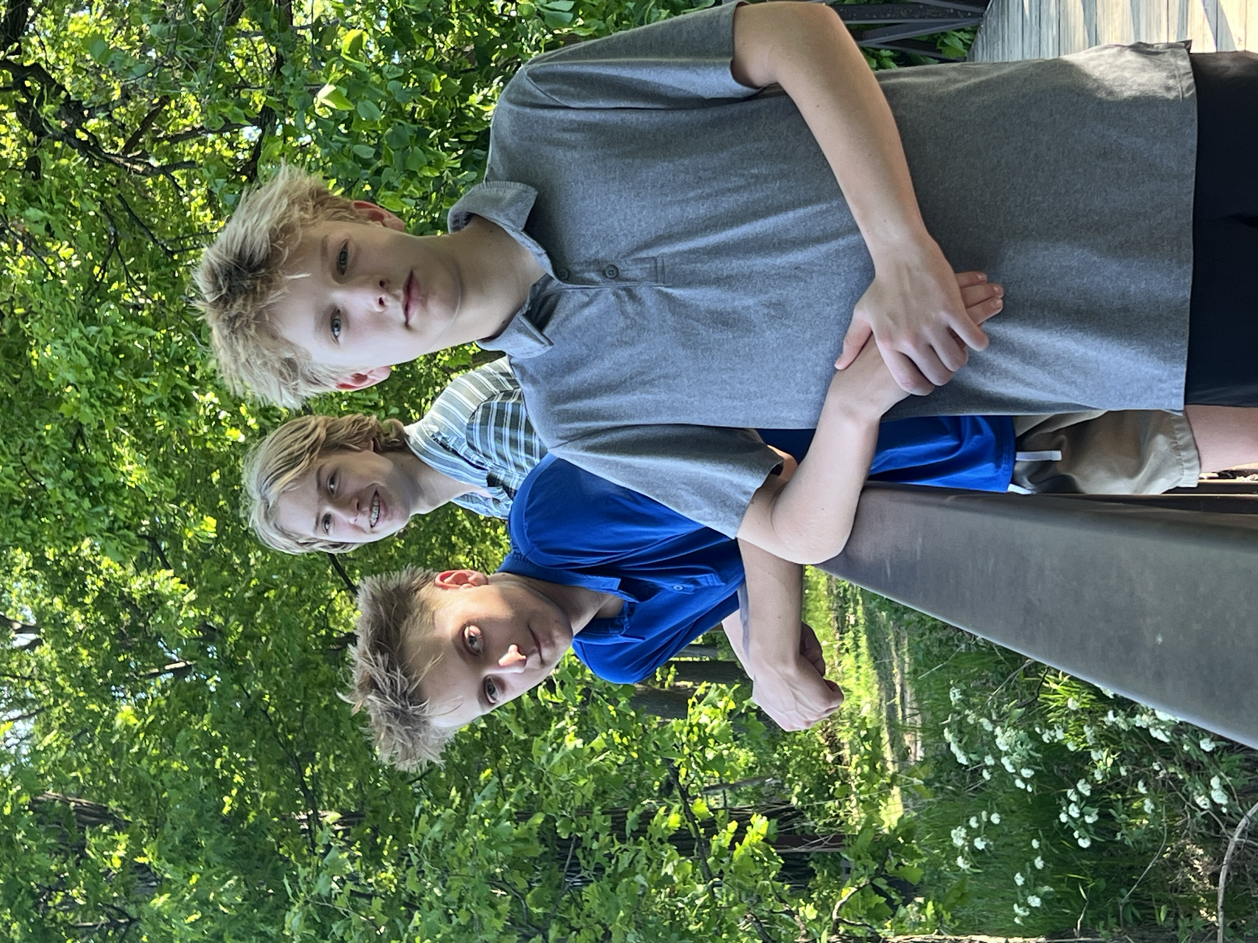 Teenage boys standing outdoors among green trees. They are casually dressed and have serious or neutral expressions. The boy in front has platinum blonde hair, wearing a gray polo shirt. The atmosphere appears to be a sunny day in a park or garden.