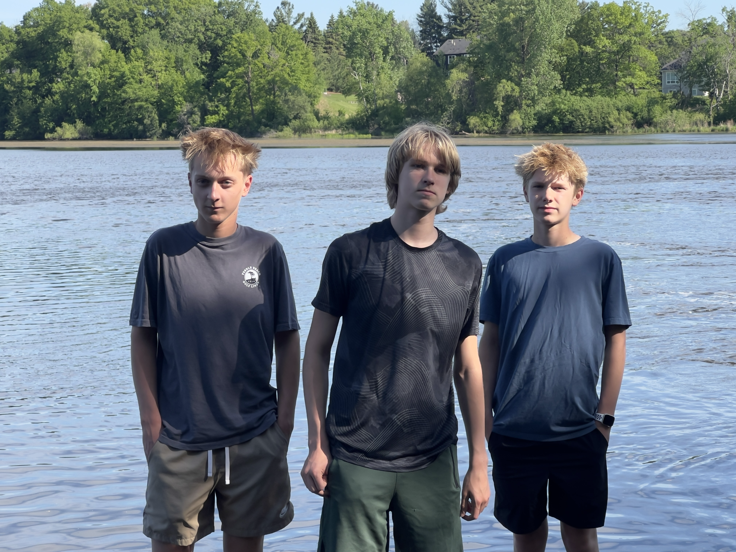 Three boys standing by a lake with green trees and houses in the background on a sunny day.