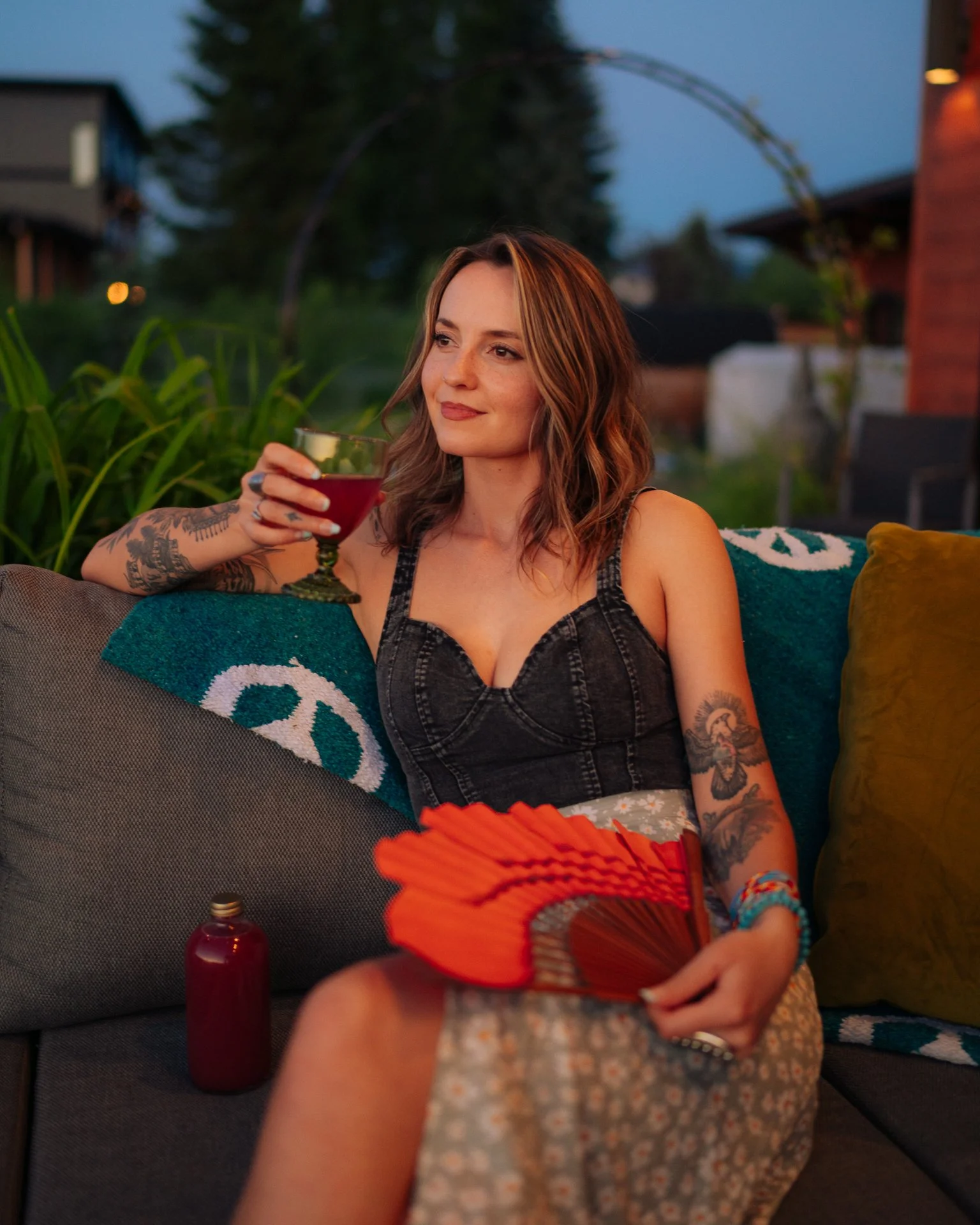 A woman with tattoos on her arm sitting on a couch outdoors during dusk, holding a red drink in a glass, with a person holding a fan in the foreground.